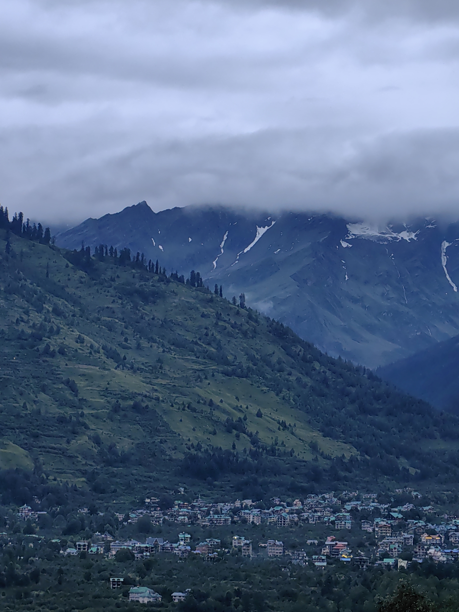The image depicts a mountainous landscape with a village nestled at the base of a steep, tree-covered hill. The village, identified as Vashisht, India, consists of numerous multi-story buildings with distinct architectural styles, many featuring green roofs, clustered together amidst dense vegetation. The hill slopes upwards, showing terraced patterns and dotted with trees. In the background, imposing mountain peaks rise, partially obscured by a thick, cloudy sky. Snow can be seen on some of the higher mountain ridges. The overall atmosphere is somber and cool, suggesting overcast or twilight conditions. There are no visible people or animals in the scene.