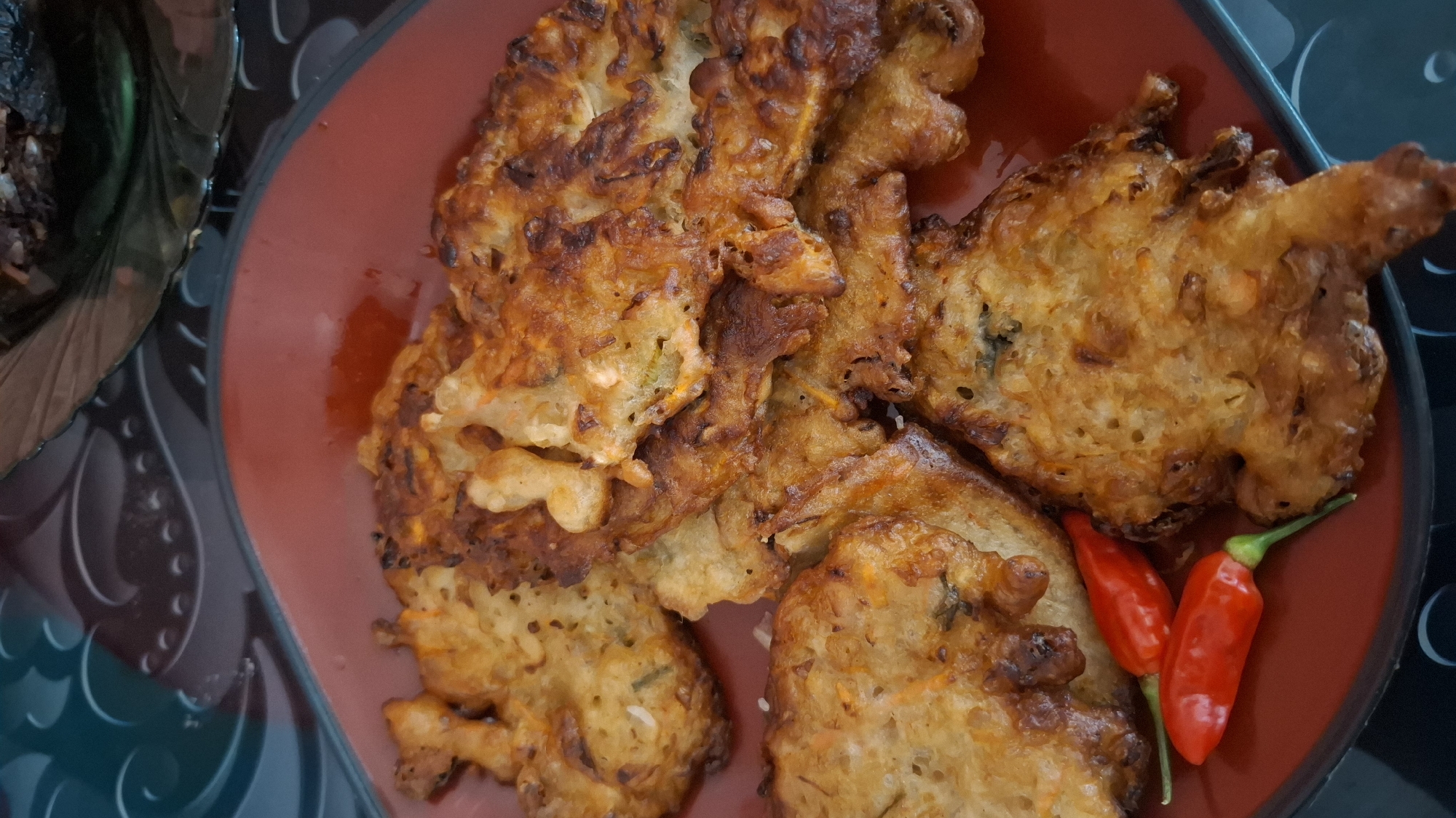 A close-up, overhead shot shows a pile of golden-brown, crispy fritters arranged on a deep red plate. Two small, bright red chili peppers are nestled in the lower left corner of the plate. The fritters appear to be made from a mixture of shredded vegetables, possibly carrots and green onions, bound together with a batter and fried to a crunchy texture. Some pieces have darker, more caramelized edges, indicating they are well-cooked. The fritters are varied in shape and size, some irregular and flat, others thicker and more bulbous. The plate itself is simple and oval-shaped, with a slight sheen. In the background, a dark, textured surface with a subtle swirling pattern is visible, and a sliver of a dark, glassy object, possibly another dish, is present in the upper right. The overall scene suggests a home-cooked meal served at a dining table. The location is stated to be Ciharashas, Indonesia.