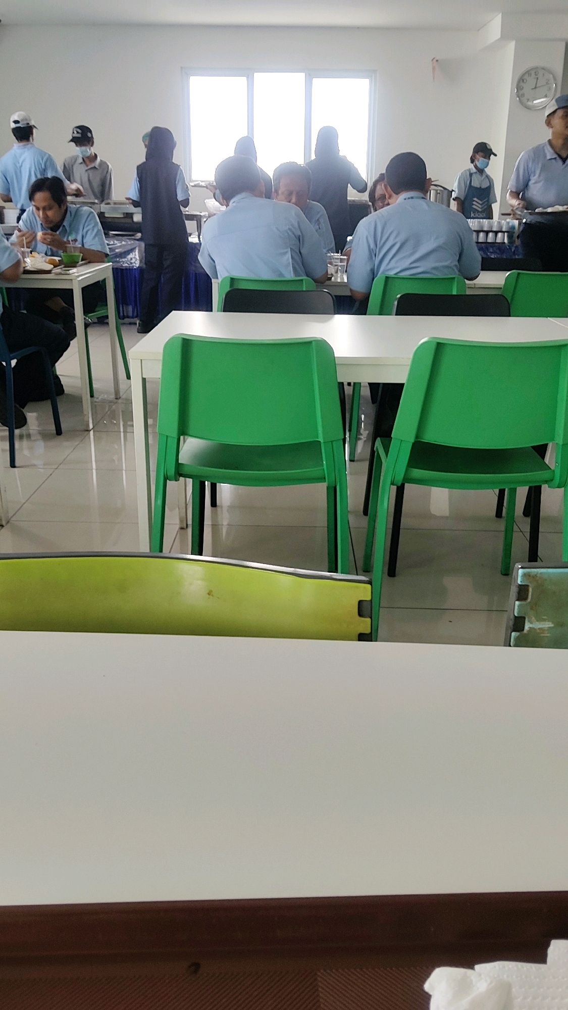 The image depicts a group of people in a brightly lit indoor cafeteria or dining hall. Several individuals, all appearing to be adults, are seated at tables, some eating. A buffet or serving station is visible in the background with people standing around it. The environment appears to be a modern facility with white walls, a tiled floor, and large windows. Green chairs are arranged around white tables in the foreground. The individuals are wearing light blue shirts and some are wearing masks or head coverings. The clock on the wall indicates it is likely daytime. This scene could be a canteen or dining area within an industrial facility or office building in Mekarmukti, Indonesia, given the context provided.