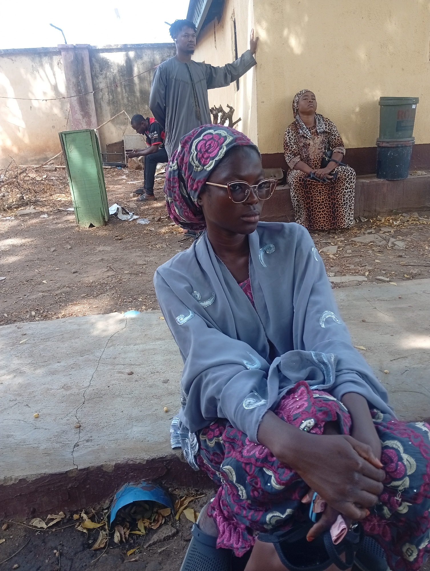 This image depicts an outdoor scene in Barden Yana, Nigeria, featuring four individuals in various states of rest or contemplation during what appears to be a sunny day.
In the foreground, a young Black woman is seated on a concrete ledge, facing slightly to the right with a thoughtful expression. She wears a grey long-sleeved robe adorned with light blue/silver embroidery, a dark headscarf with pink and black floral patterns, and light-framed glasses. Her hands are clasped in her lap, and she has light orange nail polish.
Behind her, a Black man stands leaning against a light-colored wall, wearing a grey tunic (kaftan) and looking towards the right. Further to the right, a second Black woman sits on a raised concrete platform against a light yellow painted wall. She is dressed in a leopard-print long dress and matching headscarf, with a somber or pensive expression. A black handbag rests beside her. In the background on the left, another Black man is seated on a low surface, looking down at his phone.
The ground is dusty and dry, with scattered leaves, branches, and some discarded paper/plastic. Visible objects include a green wooden panel standing upright on the left, an overturned blue bowl filled with dry leaves in the bottom left corner, and two stacked plastic buckets (a green one partially showing "ROYA" and a black one) next to the seated leopard-print clad woman. The bright sunlight casts shadows, suggesting it is mid-day. The overall scene conveys a sense of quiet observation or waiting.
