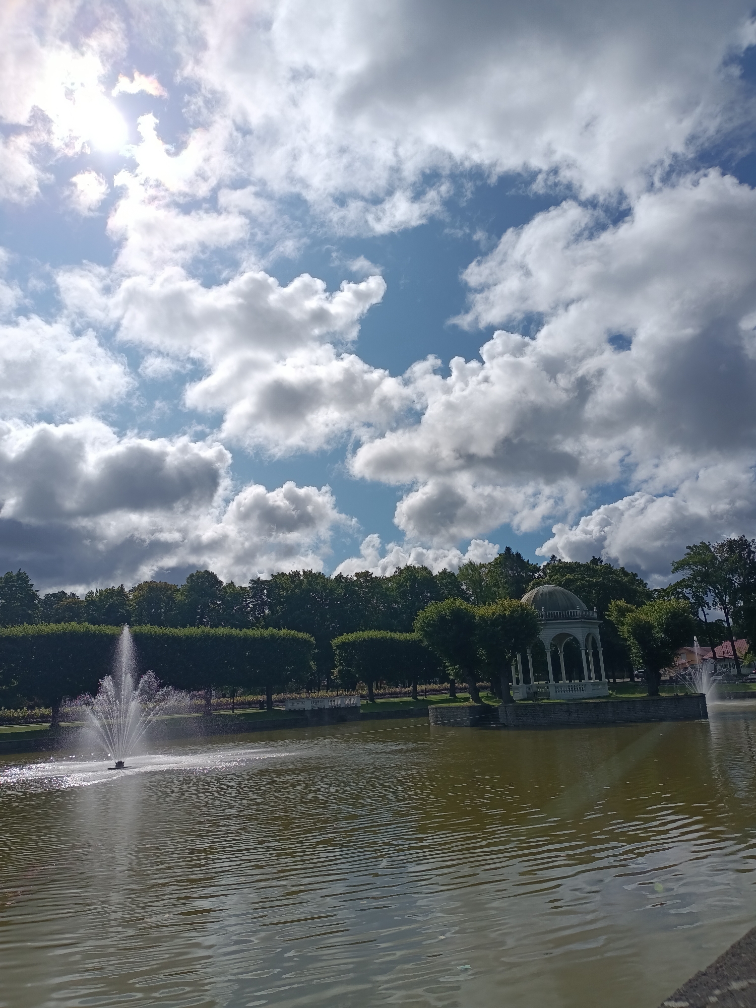 The image depicts a serene park scene with a prominent lake in the foreground. The water is calm, with subtle ripples reflecting the sunlight. A fountain sprays water upwards in the left portion of the lake. In the background, a row of lush green trees lines the shore, creating a vibrant contrast with the water. A white gazebo with a domed roof stands on a small island or peninsula within the lake, surrounded by trees. The sky above is a mix of bright blue and dramatic white clouds, with the sun partially visible and casting a bright glow. The overall impression is one of a peaceful, well-maintained park, likely a popular spot for relaxation. This scene is consistent with the landscape of Kadriorg Park in Tallinn, Estonia, known for its beautiful gardens, fountains, and historic structures such as the Kadriorg Palace and its surrounding parkland.