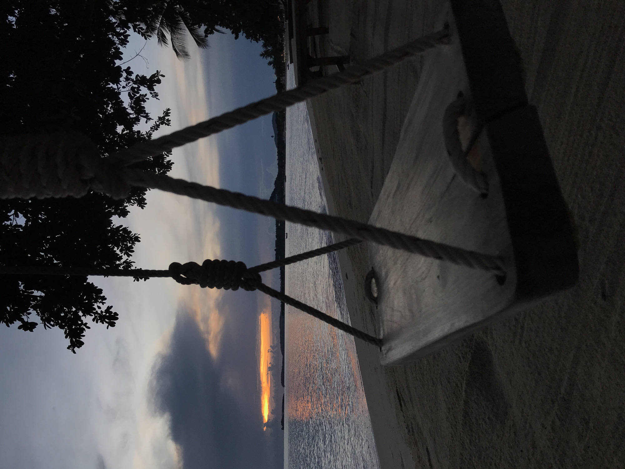 Captured at Baan Mae Nam, Thailand, this photograph features a swing set silhouetted against a dusky beachscape. The swing hangs suspended, with thick ropes tethering the wooden seat to an unseen anchor point amidst trees. A body of water reflects a sliver of bright sunset, contrasting with the surrounding dark clouds. A sandy beach stretches to the right, partially occupied by a wooden bench.