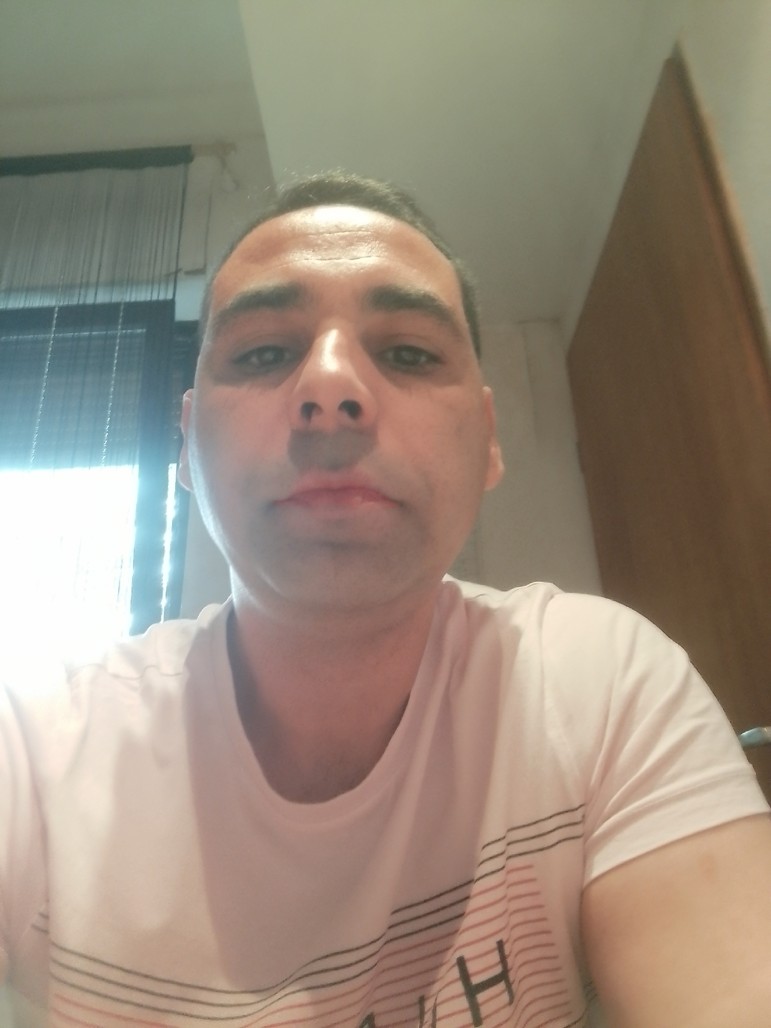 The image is a selfie taken indoors. A man with short, dark hair and fair skin is looking at the camera. He is wearing a light pink t-shirt with horizontal stripes of black, red, and white. The background is blurry and shows a window with blinds on the left and a wooden door or wall on the right. The lighting appears to be artificial, possibly from overhead lights, creating some shadows on the man's face. The scene does not contain any specific details that can be used to identify a location within the city of Delmenhorst, Germany.
