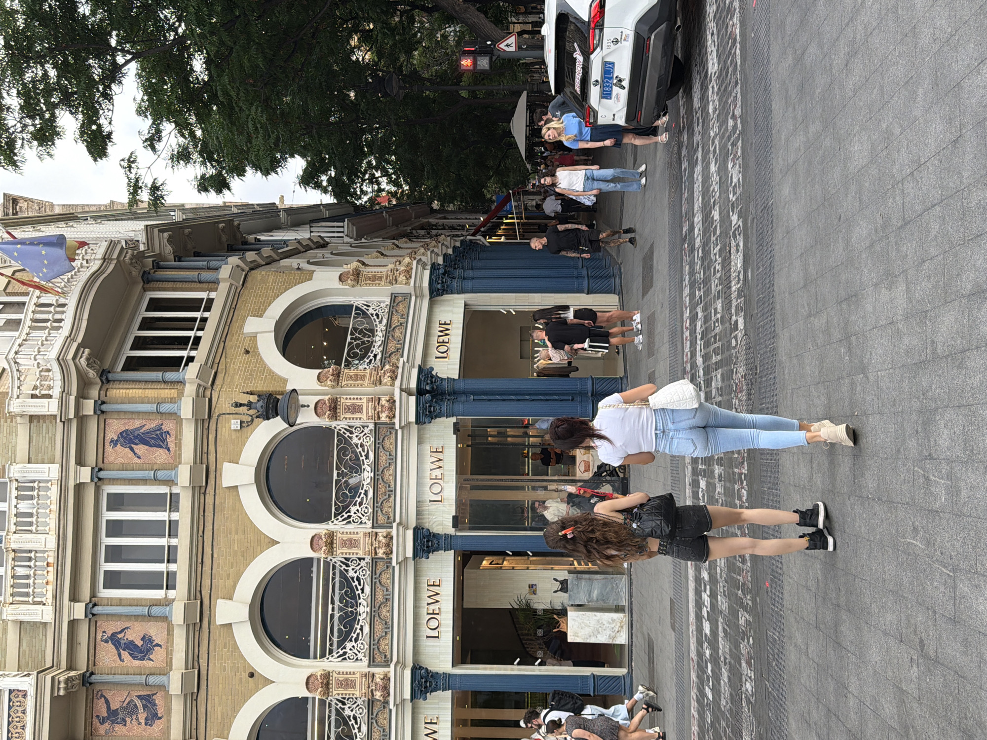 In an outdoor urban setting, a white SUV with a license plate reading "1832 LJX" is parked at the edge of a pedestrian crossing. The vehicle is facing to the right, and its rear is visible. In the background, a two-story building with an ornate facade features arched windows and the prominent "LOEWE" logo displayed across its storefront. The building's architecture is detailed, with decorative elements and blue columns. Several people are walking on the sidewalk and crossing the street. Two women are in the foreground, walking away from the camera towards the crosswalk. One is wearing denim shorts and a patterned top, and the other is wearing light blue jeans and a white t-shirt. A pedestrian signal displays a red hand. The ground is paved with cobblestones and features a patterned crosswalk. Trees line the right side of the street, and a street lamp is visible on the building's facade. The sky appears overcast.