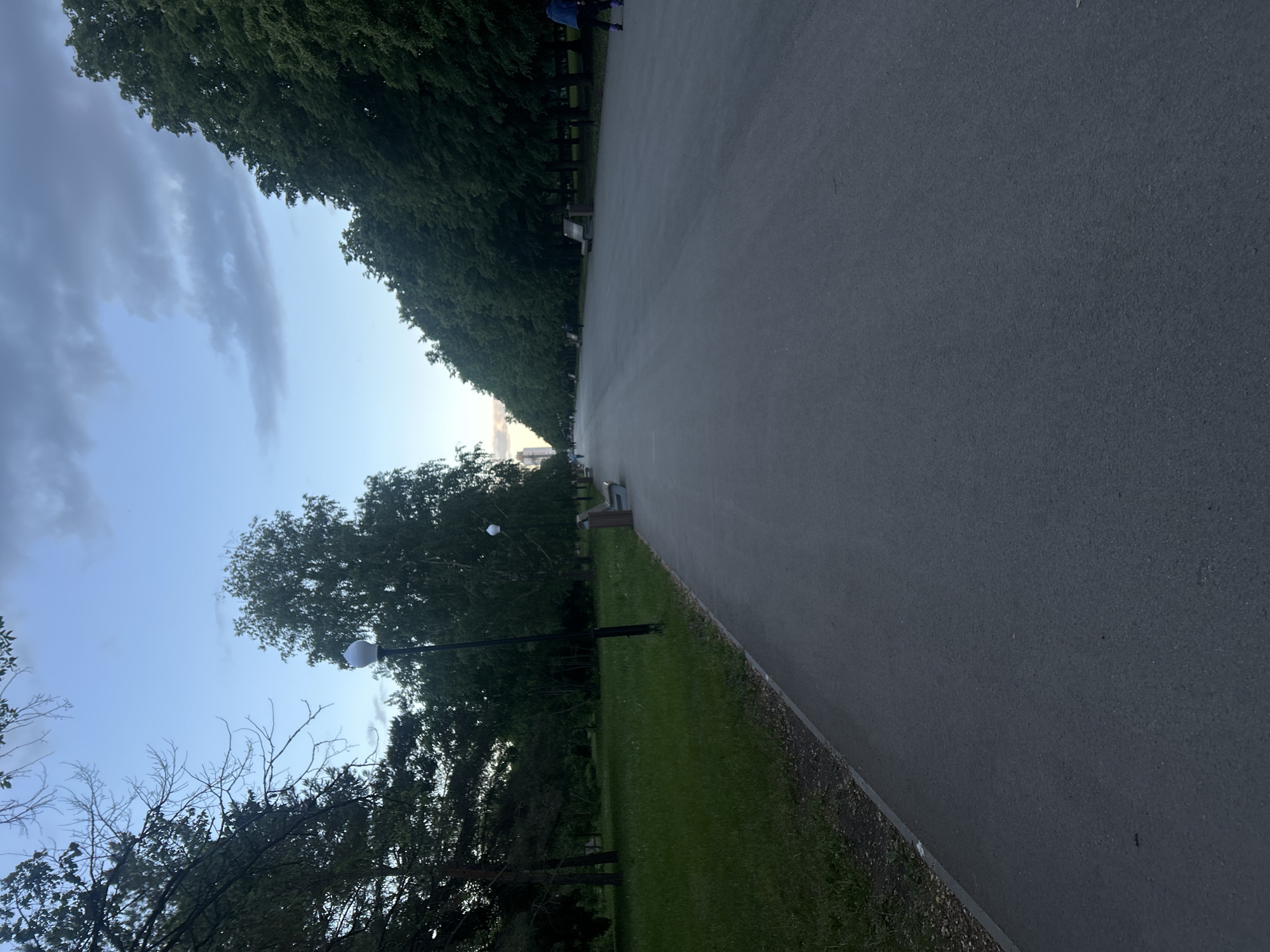 The image depicts a paved path in Tolyatti, Russia. On either side of the path are trees and grass. A cloudy sky is visible above. Street lights are placed along the path. Benches are placed periodically along the sides of the path.