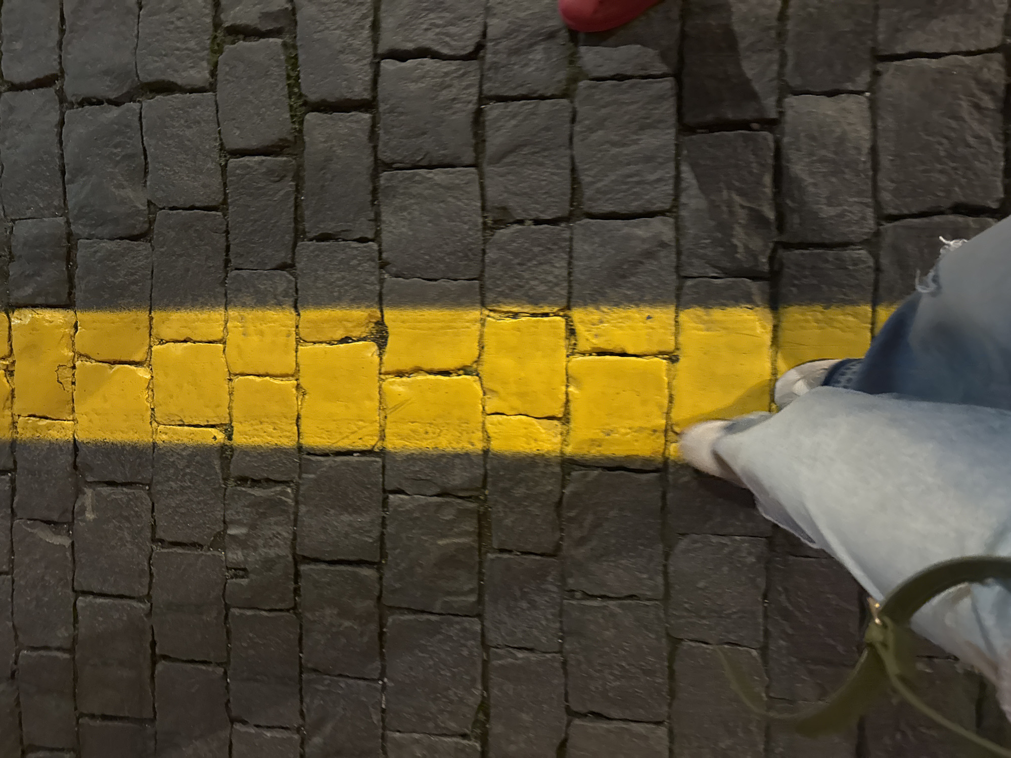 The image shows a close-up view of a cobblestone street in Moscow, Russia, with a prominent bright yellow line painted vertically down the center. The cobblestones are dark gray and appear to be worn. In the foreground, the lower legs and feet of a person wearing light blue jeans and white shoes are visible, standing just to the left of the yellow line. A dark green strap, possibly from a bag or purse, is also visible in the lower left corner. To the far right, a sliver of a red shoe is visible, suggesting another person is nearby. The lighting suggests it is either dusk or night, with artificial light casting shadows on the cobblestones.