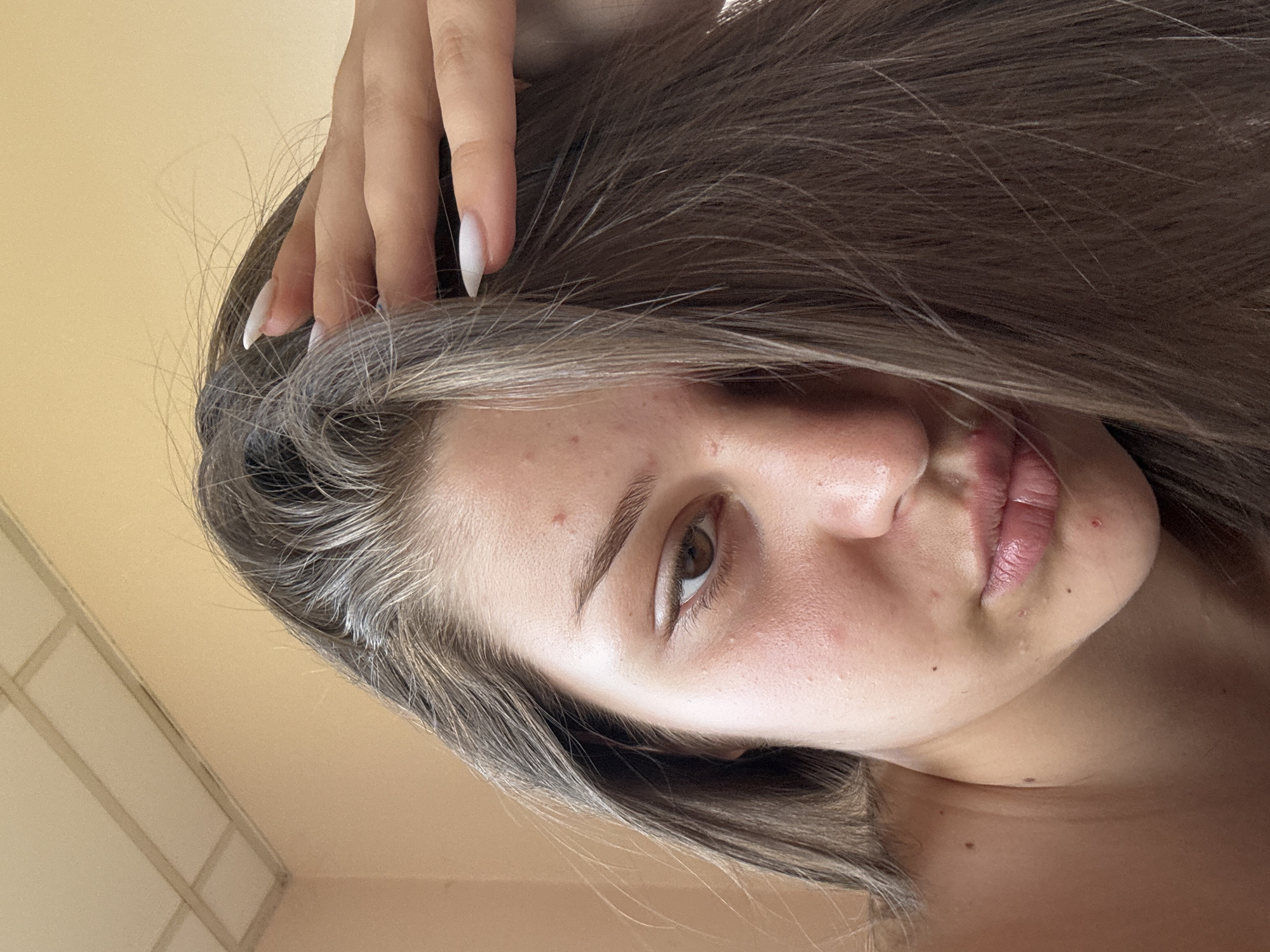 The image is a close-up, overhead, selfie-style shot of a young woman's face and upper body, positioned indoors. The background consists of a cream-colored wall and a ceiling with rectangular panels. The woman has long, dark brown hair, partially covering her face, with strands of lighter, possibly graying hair visible near her temples. Her right hand, with long, white, pointed fingernails, is placed on the left side of her head, near her temple. Her face is angled towards the camera, with her right eye, an eyebrow, part of her nose, and her lips visible. She has brown eyes and light skin with visible blemishes and some redness. Her lips are full and appear to be slightly parted and glossy. Her shoulders and neck are also visible. The lighting suggests natural light, possibly from a window.