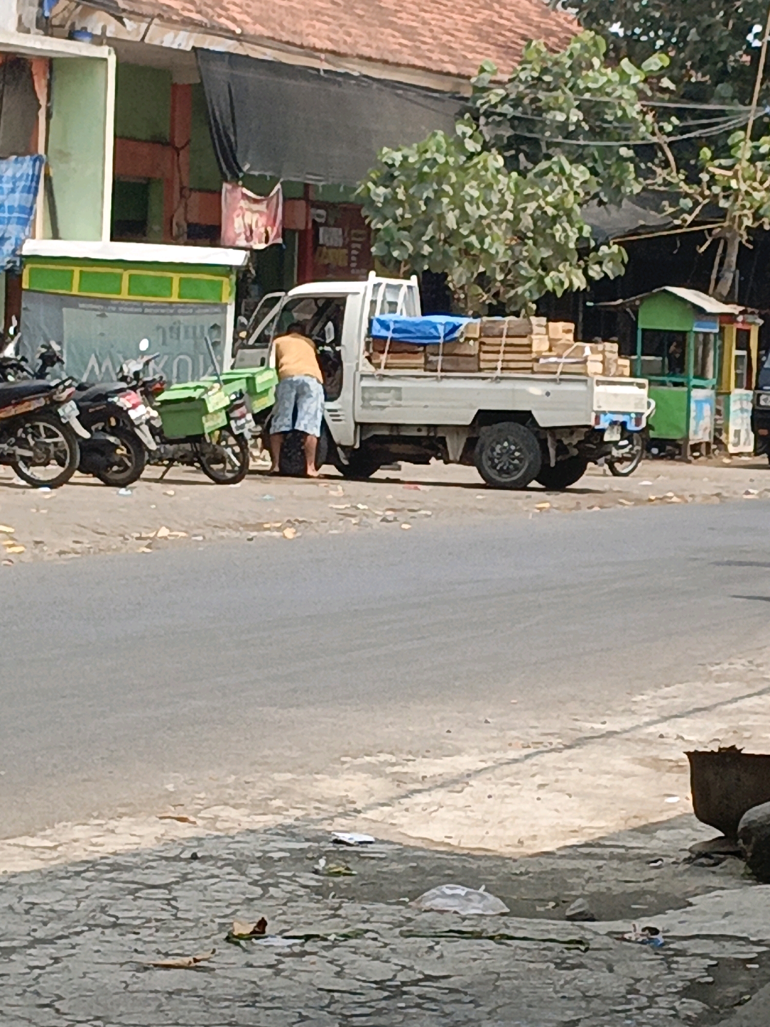 The image depicts a street scene in Betek, Indonesia, with a white pickup truck laden with wooden planks and covered with a blue tarp. A person wearing an orange shirt and patterned shorts is standing next to the truck, facing away from the camera, and appears to be interacting with the vehicle or its cargo. Several motorcycles are parked to the left of the truck. In the background, there are buildings with colorful facades, trees, and what appear to be small market stalls. The ground is paved, and there is debris scattered along the roadside. The overall impression is that of a busy street during daytime, likely in a commercial or residential area.
