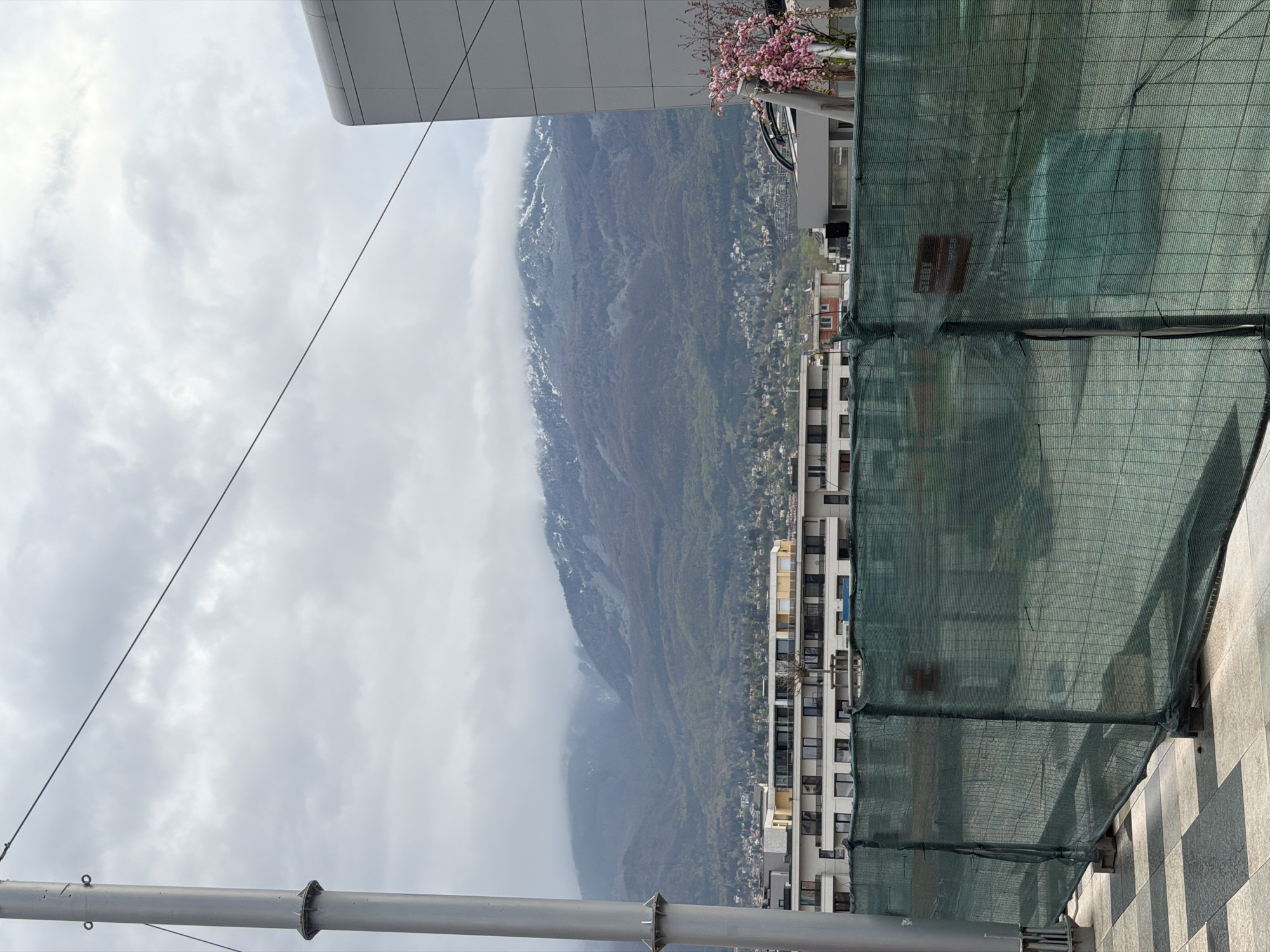 A panoramic view from an elevated position captures a city transitioning into a mountain range under an overcast sky. In the foreground, a grey and white tiled pavement is visible, alongside a tall grey metal pole on the left. A dark green mesh construction barrier stretches across the right and center-right of the frame, partially obscuring the view of buildings beyond. A small red sign with white Cyrillic text, reading 'ВНИМАНИЕ' (Attention), is visible on the green mesh.
Behind the barrier, several multi-story buildings with balconies and numerous windows form part of an urban landscape that extends towards the distant mountains. Near a modern building structure at the top right, a tree with vibrant pink blossoms adds a splash of color. The background is dominated by a large mountain range, its lower slopes covered in green vegetation, while higher elevations feature patches of snow partially enveloped by low-hanging grey clouds. The overall sky is overcast, suggesting a cloudy daytime scene. The setting appears to be a city nestled against mountains, consistent with Sofia, Bulgaria.