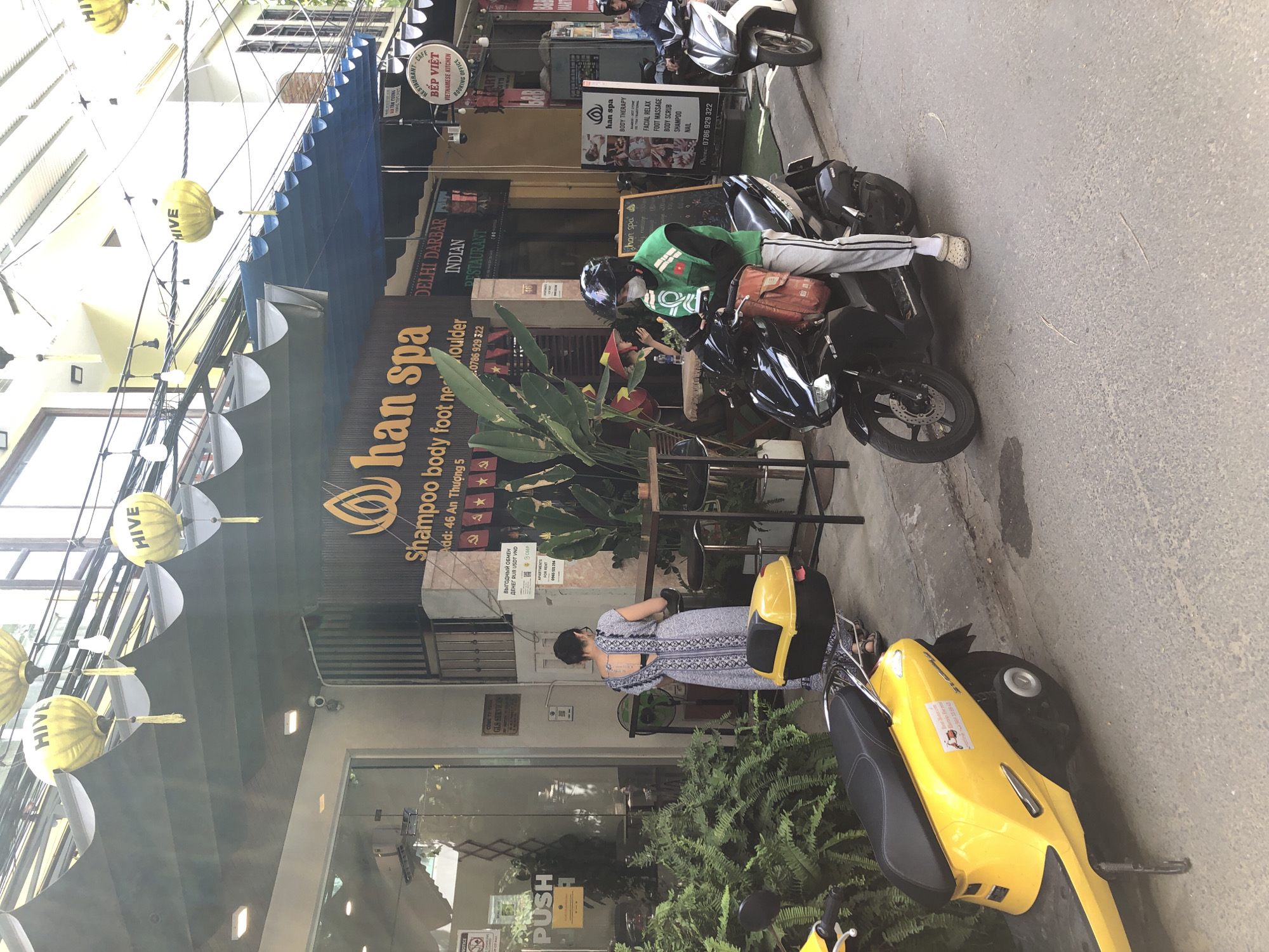 This is a street-level, medium shot photograph taken on a sunny day in Da Nang, Vietnam. The scene depicts a storefront with a blue awning extended over the sidewalk. Yellow lanterns with the word "HIVE" are hanging from wires above. The storefront features a sign for "Han Spa" offering "Shampoo Body Foot Neck Shoulder" services and lists an address: "Add: 46 An Thuong 5". To the left of the spa is a glass-fronted building with a "PUSH" sign on the door.
A woman in a patterned blue and white dress is standing on the sidewalk, facing away from the camera, near a table and chairs. In the foreground, a person wearing a green vest and a black helmet, with a white mask covering their lower face, is sitting on a black motorcycle. The person is holding a phone and has a red bag attached to the front of the motorcycle. Another motorcycle, white with black accents, is parked to the right. A yellow scooter is parked on the left side of the street, partially visible. The street is paved with asphalt. Lush green foliage is visible in front of the spa entrance.