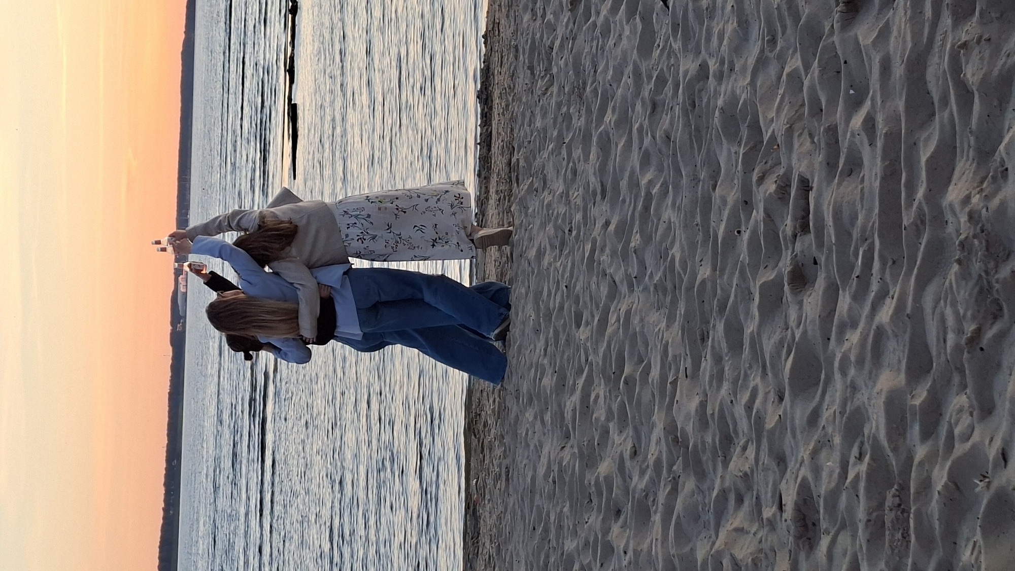 Three individuals stand on a sandy beach, facing a body of water with a calm surface. The sky above is a gradient of orange and pink, indicating either sunrise or sunset. Two of the individuals, positioned in the foreground and center, are holding up bottles, seemingly in a toast. They are embracing each other, with one person wearing a light-colored jacket over a floral dress, and the other in a blue blazer. The third individual, partially obscured, is on the left, also embracing the person in the blue blazer. The background shows a faint outline of land with some structures. The sandy beach in the foreground has ripples and indentations, suggesting a natural, possibly undisturbed, environment. The scene evokes a sense of friendship and celebration at dusk or dawn by the sea.