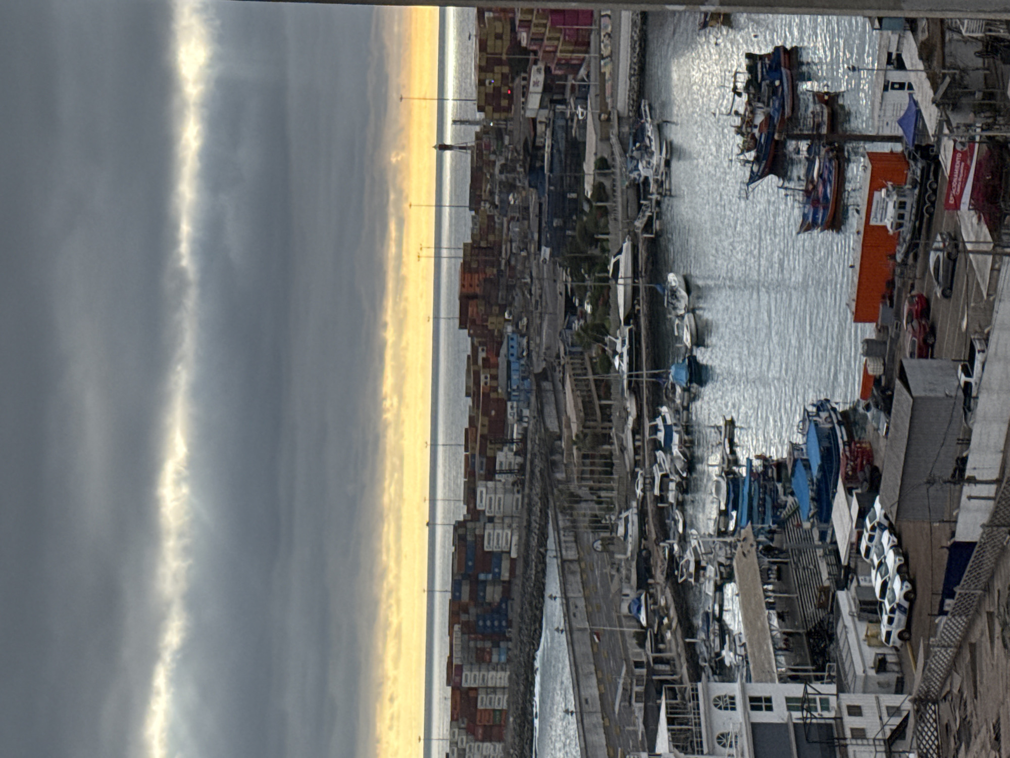 This is a high-angle, eye-level view of the port area of Iquique, Chile, under a cloudy sky with a bright yellow-orange band of sunlight near the horizon. The scene is divided into distinct sections: the sea with a breakwater and stacked shipping containers in the background, and a marina and port facilities in the foreground and midground.
In the background, the sea is calm, meeting the land at a fortified breakwater lined with numerous stacked shipping containers of various colors, including red, white, and blue. Tall light poles are visible throughout this area. Further in the distance, a lighthouse can be seen.
The midground and foreground are dominated by a bustling marina and port infrastructure. Numerous boats of various sizes are docked in the water. These include small fishing boats, some with blue tarps, and larger pleasure crafts. A prominent white building with multiple windows and a covered walkway with stairs leading down to the water is situated on the left side of the image. Adjacent to this building, several white utility vehicles, possibly police or security, are parked in a row on a paved area. To the right of these vehicles, a large orange shipping container is visible, as well as a smaller blue tent structure.
Further to the right, several more fishing boats are moored. The water in the marina reflects the sky and the boats, creating a shimmering effect. The overall impression is one of an active working port with a mix of commercial and recreational maritime activity. The lighting suggests it is either early morning or late afternoon.