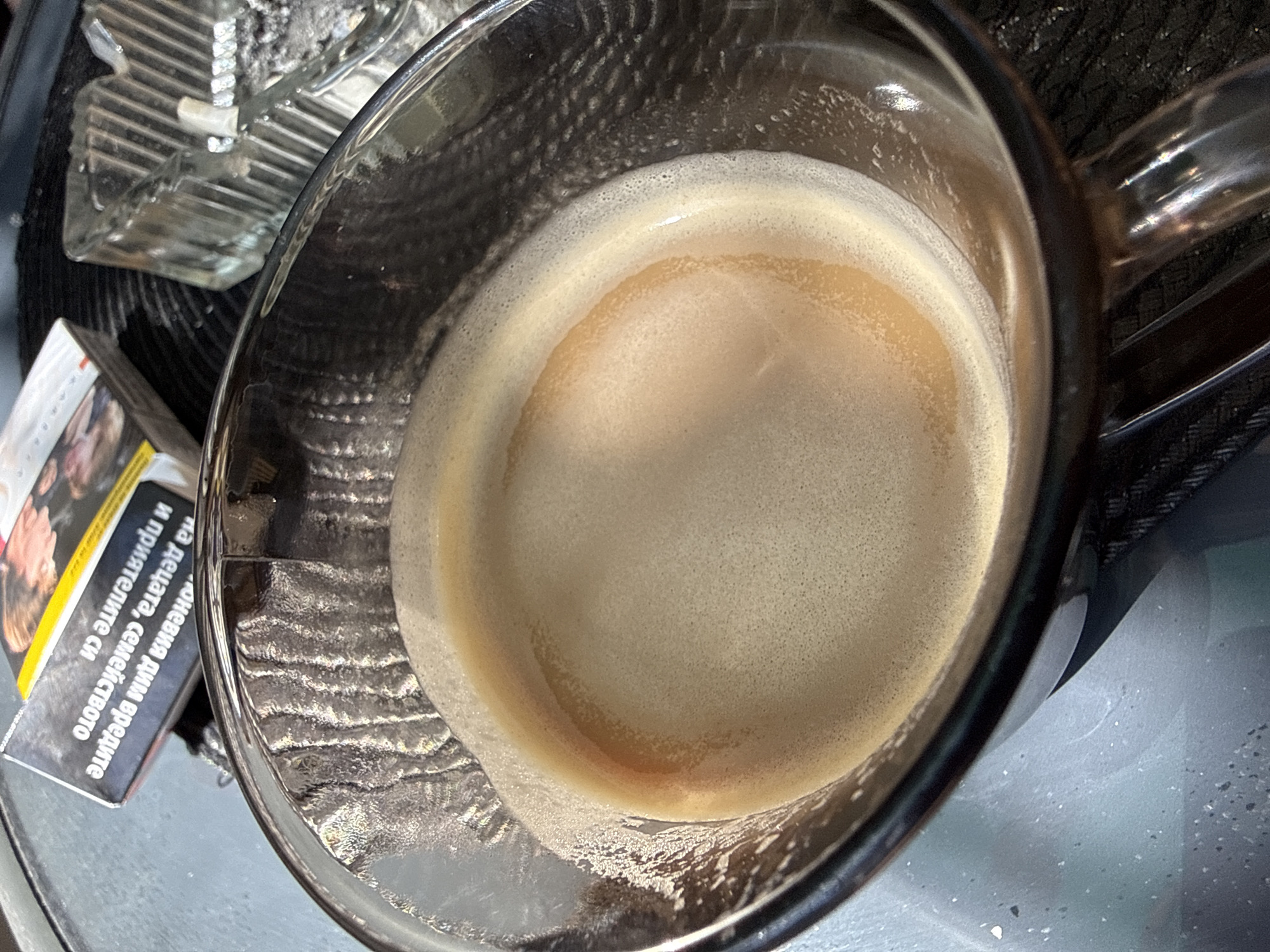 A high-angle shot depicts a clear glass mug filled with espresso, presenting a substantial layer of crema. The mug is placed on a surface with a greyish, slightly mottled appearance. To the left of the mug, a partially visible pack of cigarettes rests, positioned near a clear glass ashtray containing ashes. The scene evokes an intimate setting, likely a cafe or personal space. No people are visible. The location in the city of Lovech, Bulgaria is not confirmed from the visual data.
