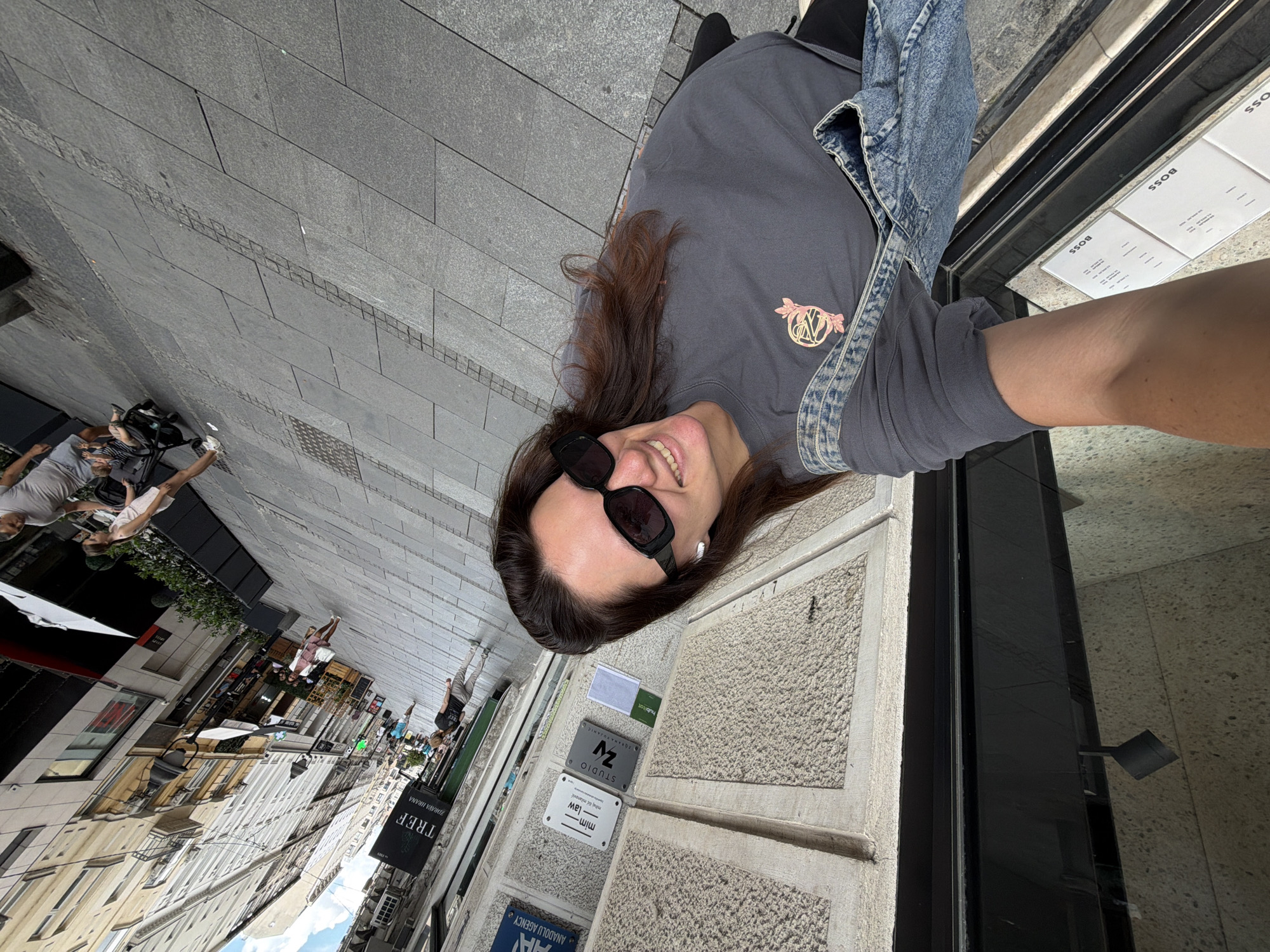This is a portrait-oriented, candid, outdoor photograph taken from a low angle, capturing a young woman with long, brown hair and dark sunglasses. She is smiling directly at the camera, wearing a dark gray t-shirt with a logo and a denim jacket over it. The photo appears to be taken in an urban setting, likely on a sidewalk lined with shops and buildings. The pavement is made of gray stone tiles. In the background, there are various signs, including one for "TREF ZDRAVA HRANA" (TREF HEALTHY FOOD), and others that are partially visible or blurred. The overall impression is that of a casual day out in a European city. The lighting suggests it is daytime, likely a sunny day given the shadows and brightness. The woman's expression is cheerful and relaxed. The presence of shops and the urban architecture suggest a commercial or pedestrian area.