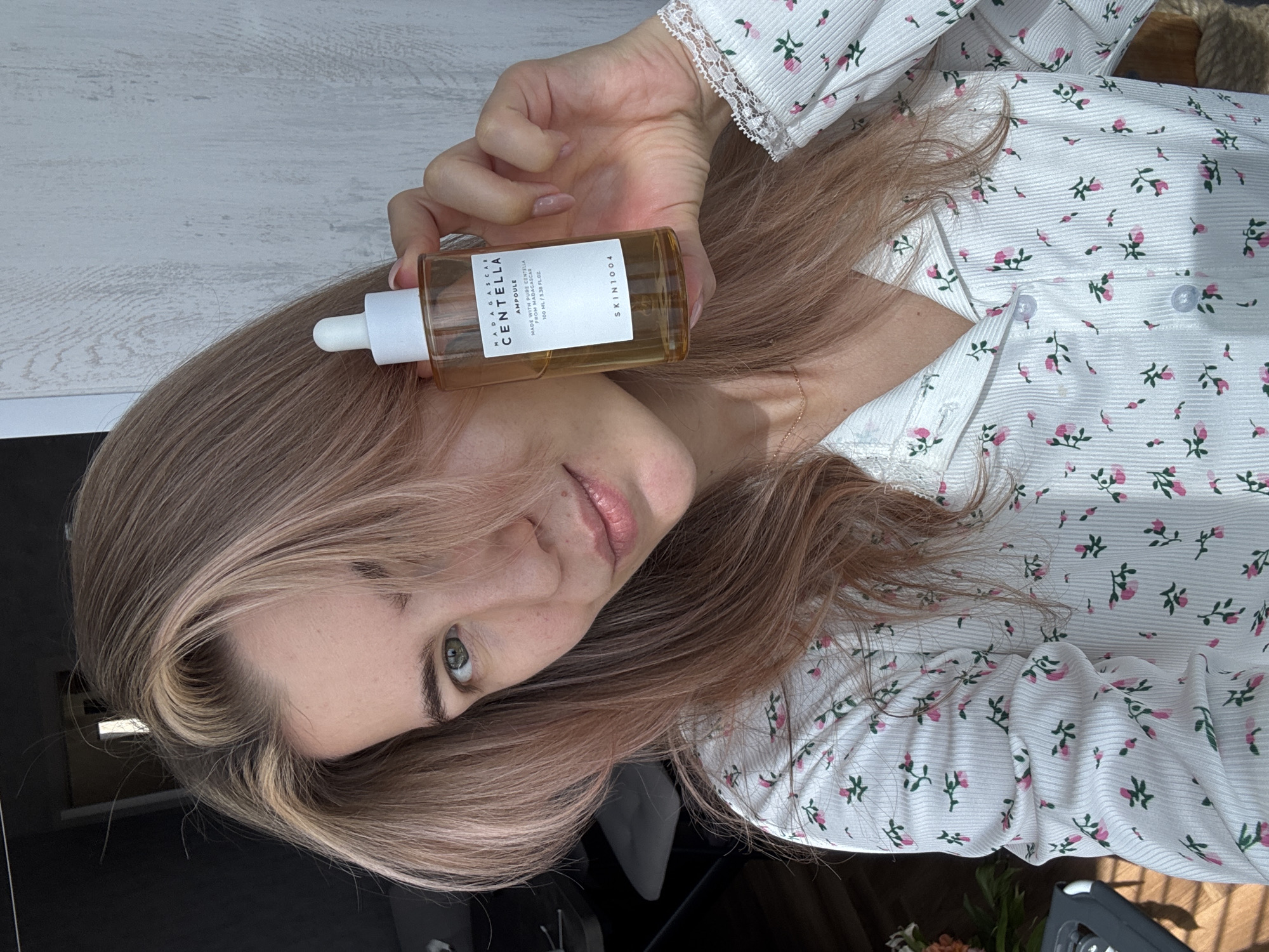 A woman is holding a bottle of Skin1004 Madagascar Centella Ampoule in her right hand. She is wearing a white shirt with pink floral patterns. Her hair is long and light brown with pink undertones. The background appears to be a light-colored surface, possibly a table or floor. The image is rotated. The location is unknown.