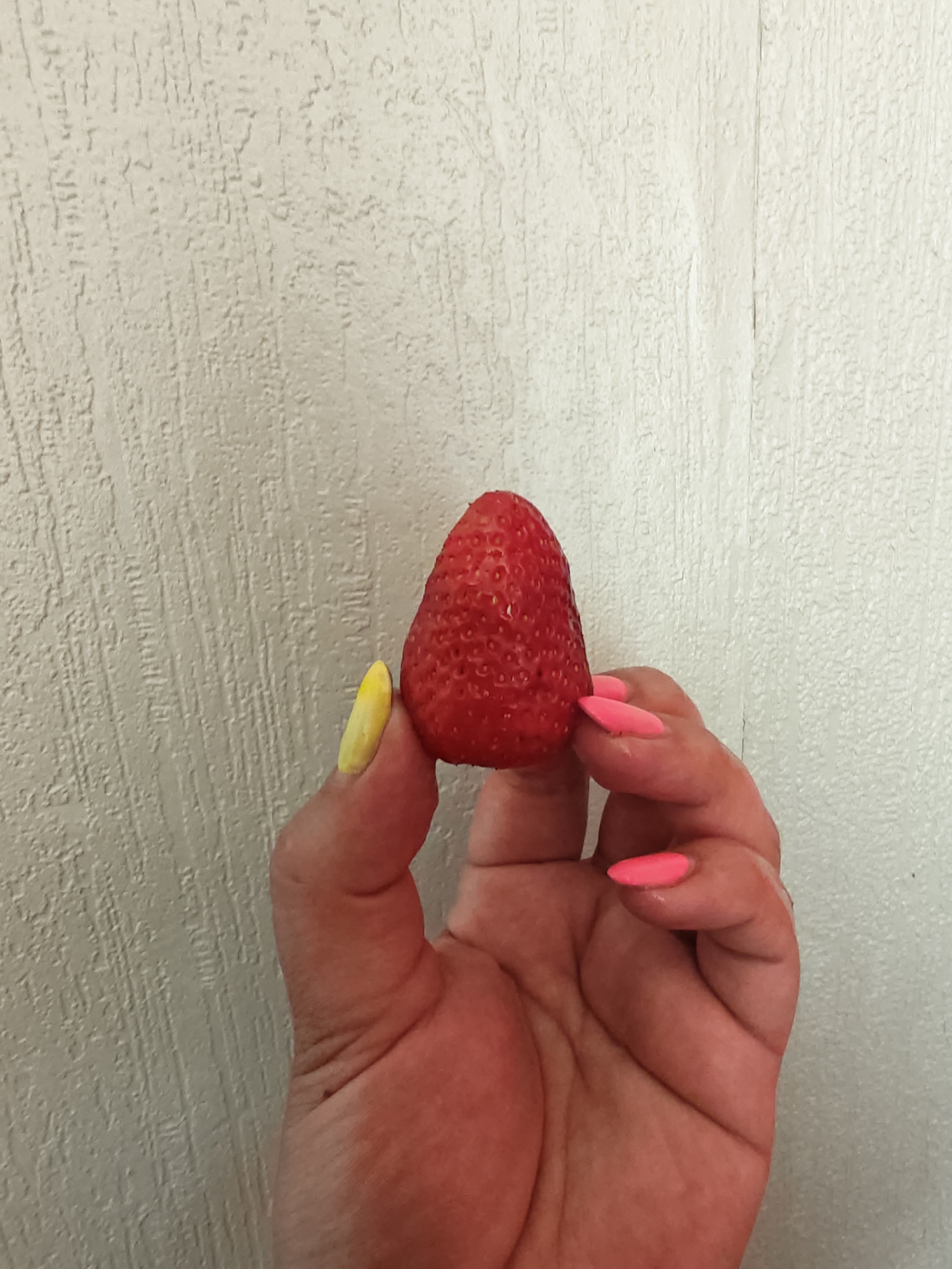 This close-up, vertical shot features a human hand holding a single, vibrant red strawberry against a textured, light-colored wall, set in Chaikovsky, Russia. The ripe strawberry is a deep red, with its small seeds (achenes) clearly visible on its surface, and appears to have a few tiny water droplets, suggesting freshness. The hand displays a striking manicure: the thumb is painted a bright yellow, while the other visible fingernails on the index, middle, ring, and pinky fingers are a bold neon pink. The background is an off-white or light beige wall, characterized by a distinct vertical, textured pattern, possibly from wallpaper or a specific painting technique. A faint vertical seam or junction is discernible on the right side of the background. The scene is evenly and brightly lit, likely indoors during the daytime, and no text is visible. The image focuses on the fruit and the hand, conveying a simple, direct presentation of the strawberry.