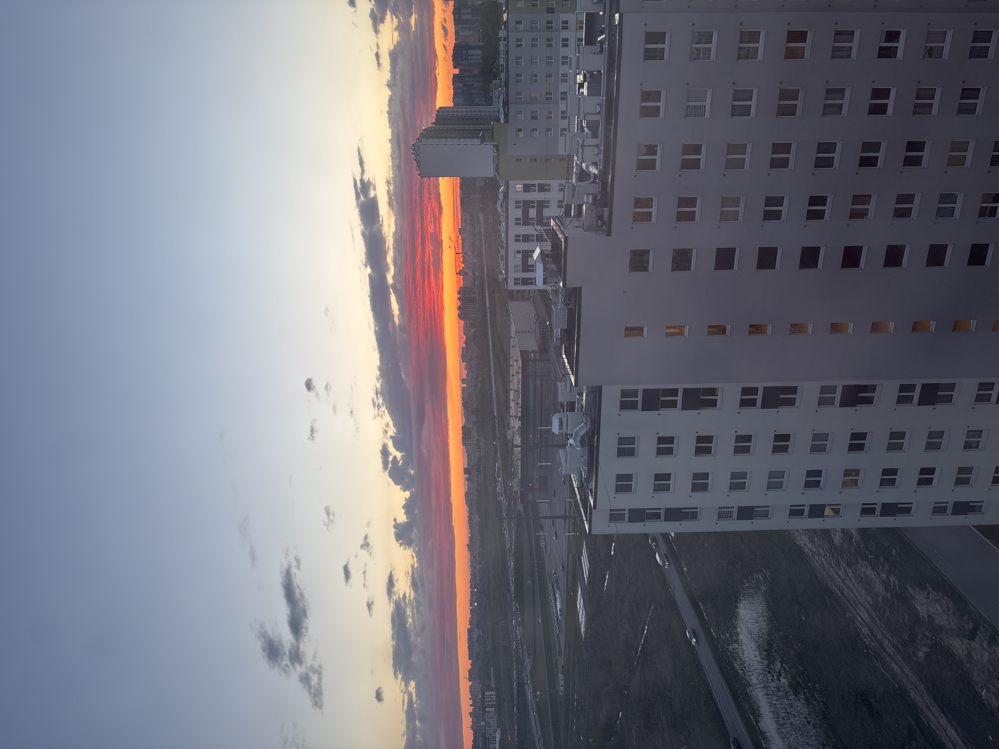 The image depicts a sunset over Vsevolozhsk, Russia, as viewed from a high vantage point. The sky transitions from blue to orange and red near the horizon, with dark clouds interspersed. Several multi-story buildings, likely apartment complexes, are visible, with numerous windows suggesting residential occupancy. The foreground reveals roadways and other urban infrastructure, possibly including train tracks. No people are clearly discernible.