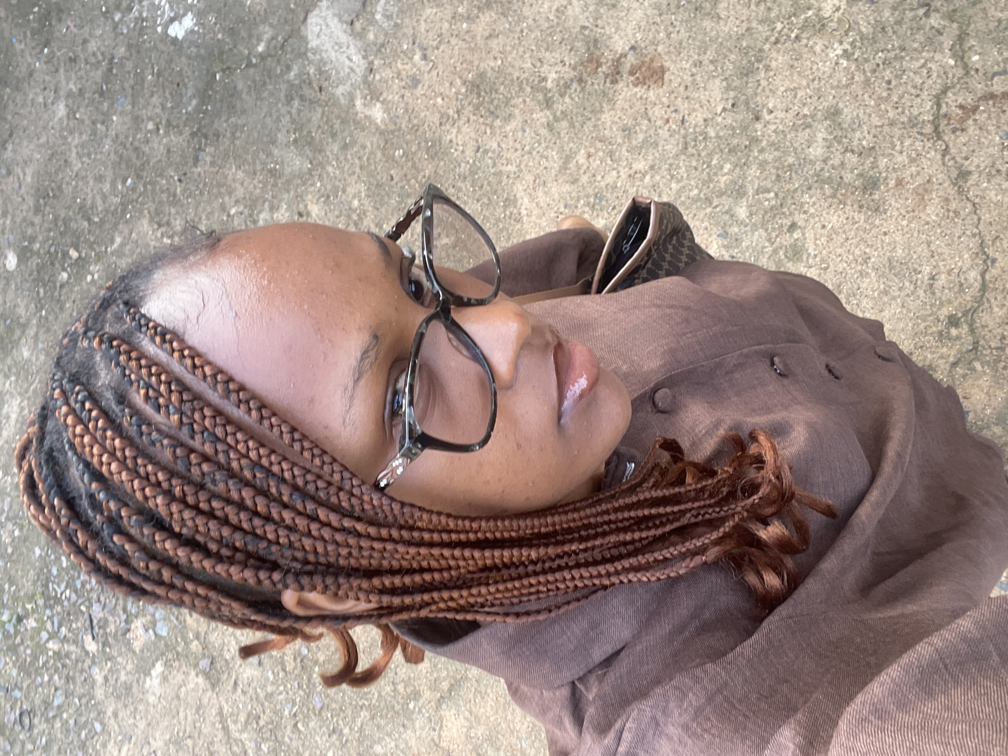 The image shows a close-up, overhead shot of a young Black woman. She is wearing a brown linen shirt and glasses. Her hair is styled in braids, some of which fall over her shoulder. She is looking upwards and to the right, with a neutral expression. The background appears to be a concrete surface, possibly a sidewalk or paved area. The lighting suggests it is daytime. The image was taken in Rafin Par, Nigeria.