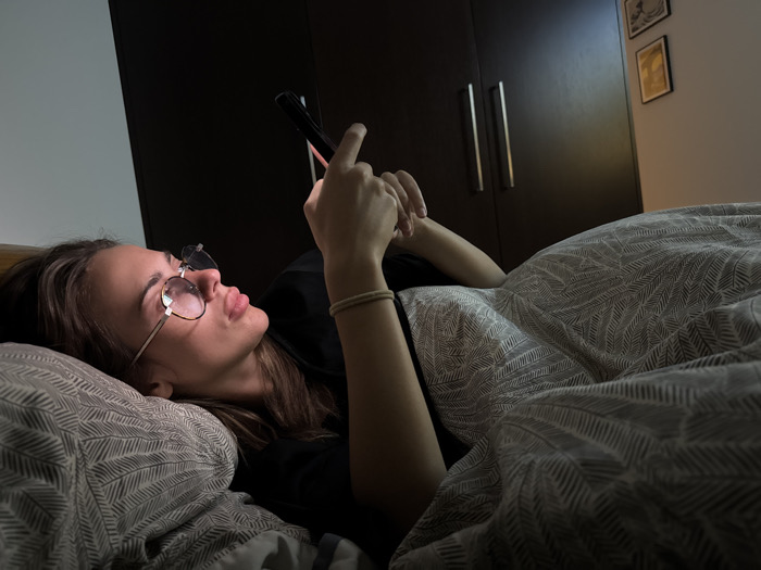 A person lies in bed, partially under a gray-patterned blanket, illuminated by the glow of a handheld phone. The person wears glasses and is propped up slightly on a patterned pillow. Behind the bed, a dark brown wardrobe with silver handles is visible against a partially lit wall, adorned with two framed pictures. The overall atmosphere is dimly lit and intimate.