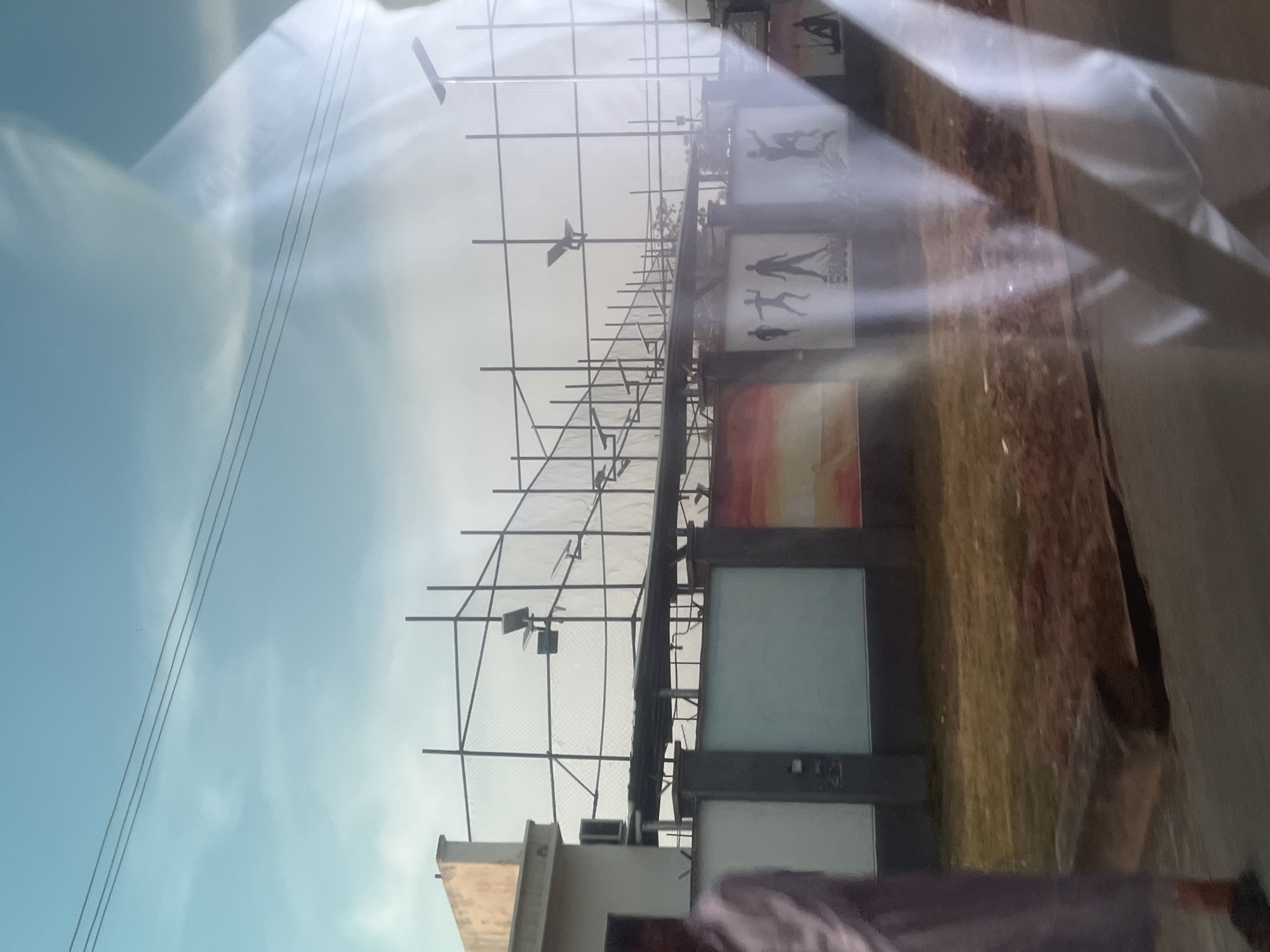 The image depicts an outdoor scene, likely a sports facility or recreational area, located in Jalingo, Nigeria. The foreground is dominated by a reflection on a glass surface, obscuring parts of the scene. Beyond the reflection, a fence constructed of metal poles and netting encloses an area with scaffolding. Several athletic figures are depicted in silhouette on posters attached to the fence. The sky above is a pale blue with scattered clouds. A person in a light-colored garment is partially visible on the left side of the image, appearing to be walking. The ground appears to be a mix of dry grass and soil.