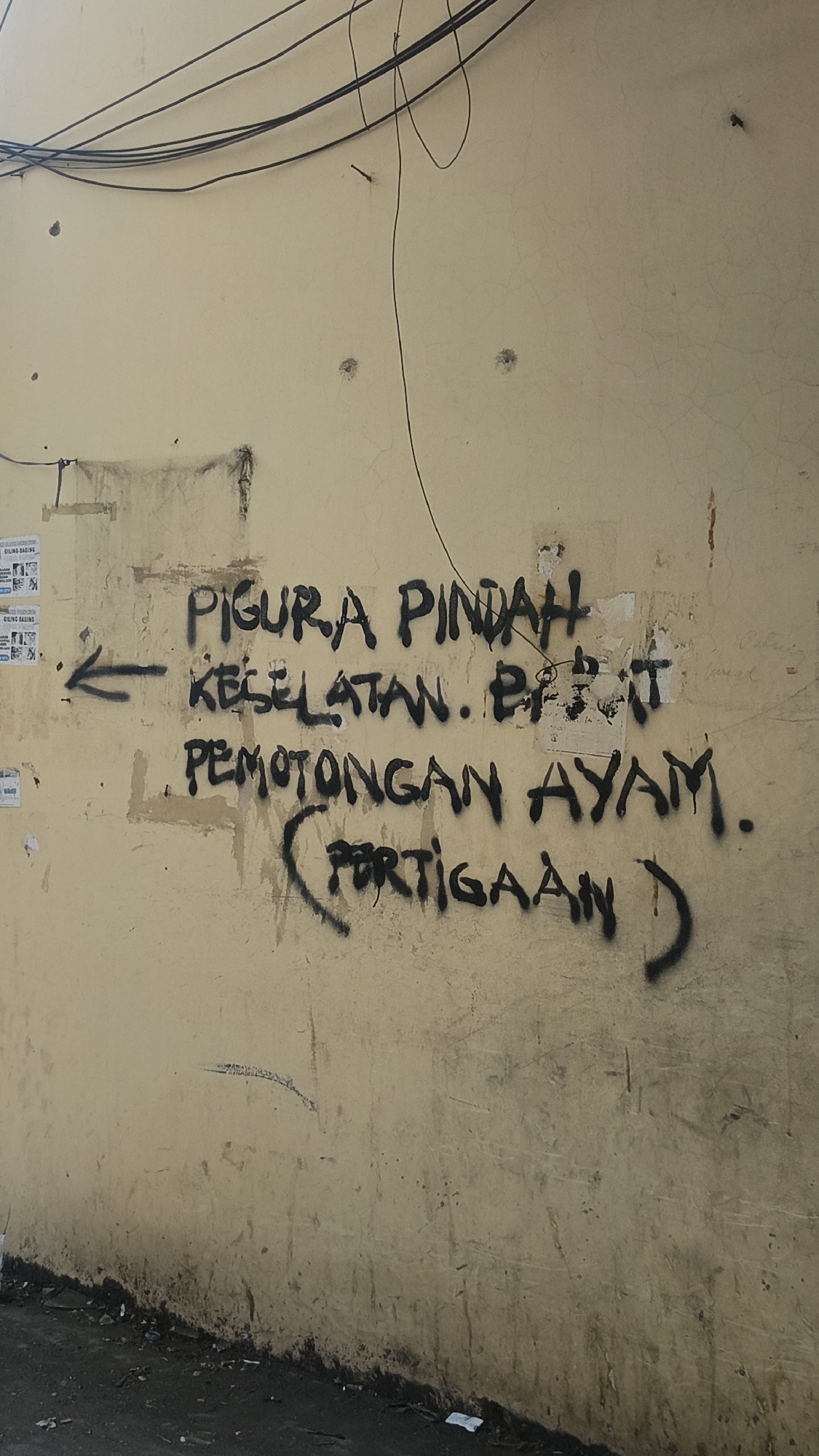 The image captures a close-up of a weathered, light-colored wall in Jekulo, Indonesia, covered in black spray-painted graffiti. The graffiti appears to be in Indonesian and reads: "FIGURA PINDAH KESELATAN. POTONGAN AYAM. (PORTIGAAN)". An arrow points to the left, preceding the text. The wall itself shows signs of age and damage, with stains, cracks, and some peeling paint. Overhead, a tangle of black electrical wires is visible against the wall. The bottom portion of the image reveals the dark, uneven ground, littered with debris, suggesting an outdoor urban or semi-urban environment.