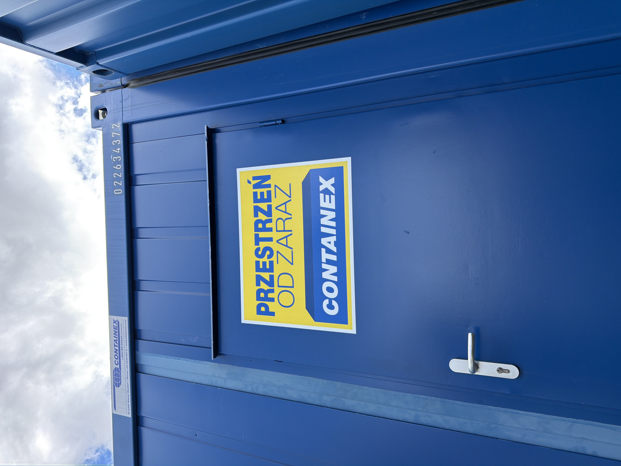 The image shows the side of a blue shipping container. A white sign with yellow lettering is affixed to the container door. The sign reads "PRZESTRZEN OD ZARAZ" at the top and "CONTAINEX" at the bottom. The container has a silver door handle and a lock. In the background, there is a cloudy sky. The container is located in Stróża, Poland.