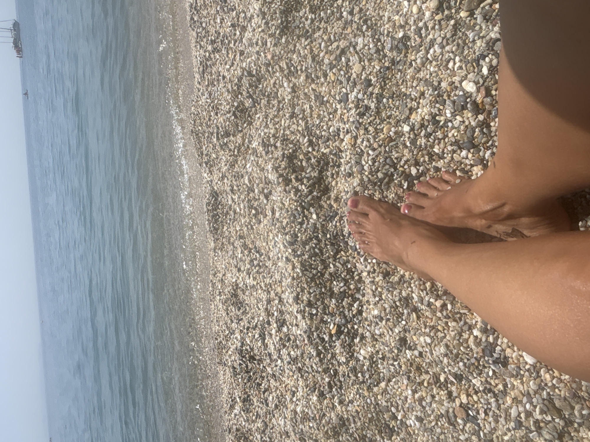 This is a first-person perspective shot of a person's legs and feet on a pebble beach in El Parador de las Hortichuelas, Spain. The ocean is visible in the background with a few small boats on the horizon. The water is a light blue color and appears calm. The beach is covered in small, multi-colored pebbles. The person's feet are visible in the foreground, with painted toenails. The lighting suggests it is daytime.