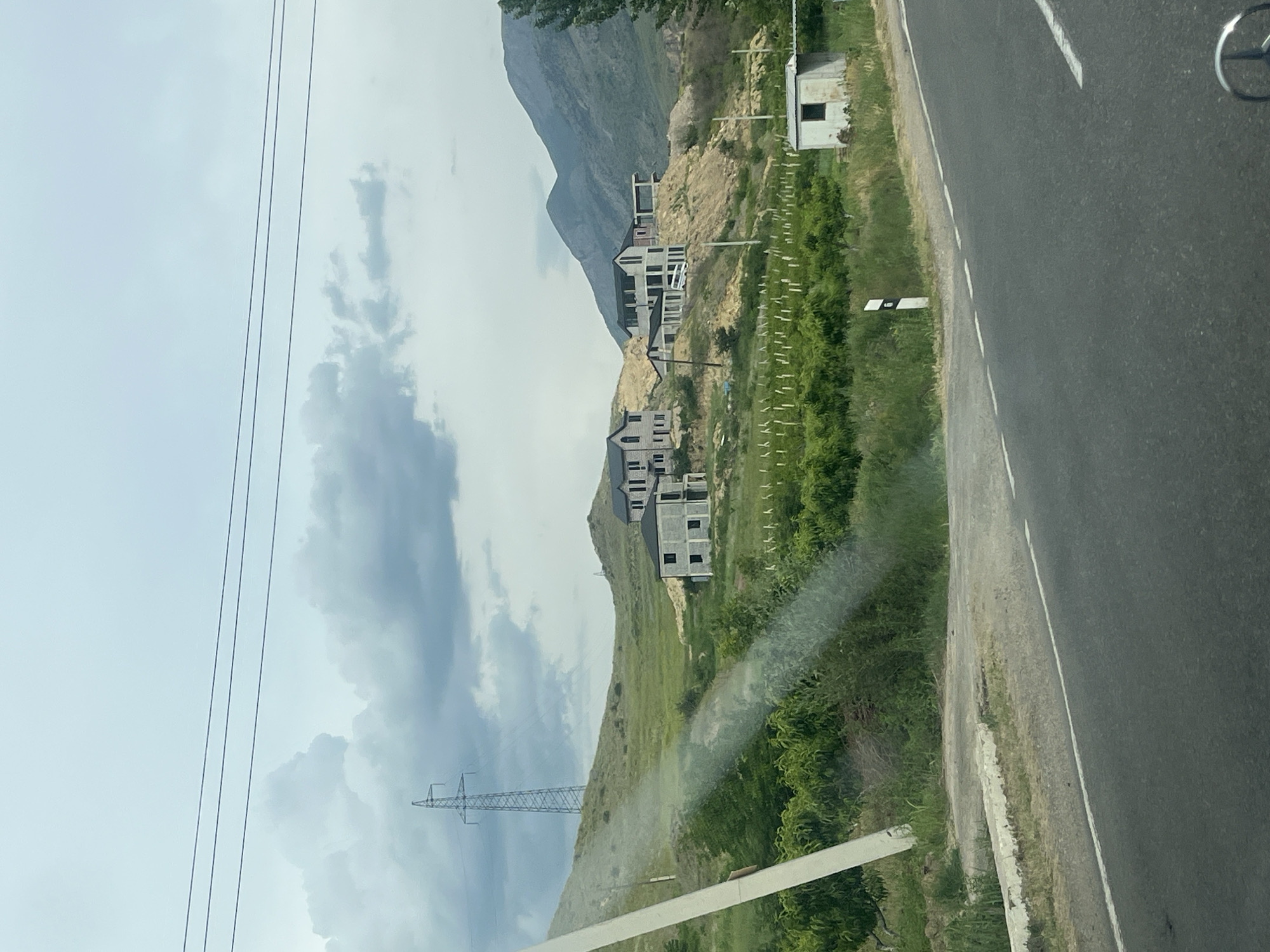 The image, taken from inside a vehicle, captures a mountainous landscape alongside a road. The perspective is from the front passenger seat, with a partial view of a Mercedes-Benz steering wheel emblem visible in the upper right corner.
On the right side of the frame, a two-lane asphalt road runs parallel to the viewer's direction, marked by a solid white line on its right edge and a broken white line on its left, separating the lanes. A narrow dirt shoulder borders the road, followed by a strip of green grass and bushes. A small, white, rectangular building or shack with a single window is situated near the road on a slight incline. Further along, a small, white roadside marker is visible.
To the left of the road, the terrain rises into steep hills and mountains. The foreground and midground show slopes covered in green grass and scattered bushes. Several unfinished concrete and block-built houses or structures are under construction on these slopes, appearing to be multi-story and varying in size. Some have visible scaffolding or exposed rebar, indicating ongoing development. A distinct pattern of small, upright wooden stakes or posts can be seen in rows on the grassy areas between the houses, possibly indicating vineyard plantings or property divisions.
In the mid-distance and background, larger mountains with rugged, rocky faces rise prominently under a cloudy sky. The sky itself is overcast, with patches of bright white and darker grey clouds. Power lines stretch across the upper left portion of the sky, leading to a tall metal lattice transmission tower visible on a lower hill to the left.
The scene depicts a daytime setting in what appears to be a rural or semi-rural area undergoing development, likely in Chiva, Armenia, given the context. No specific human activity is discernible, and no legible text is visible.