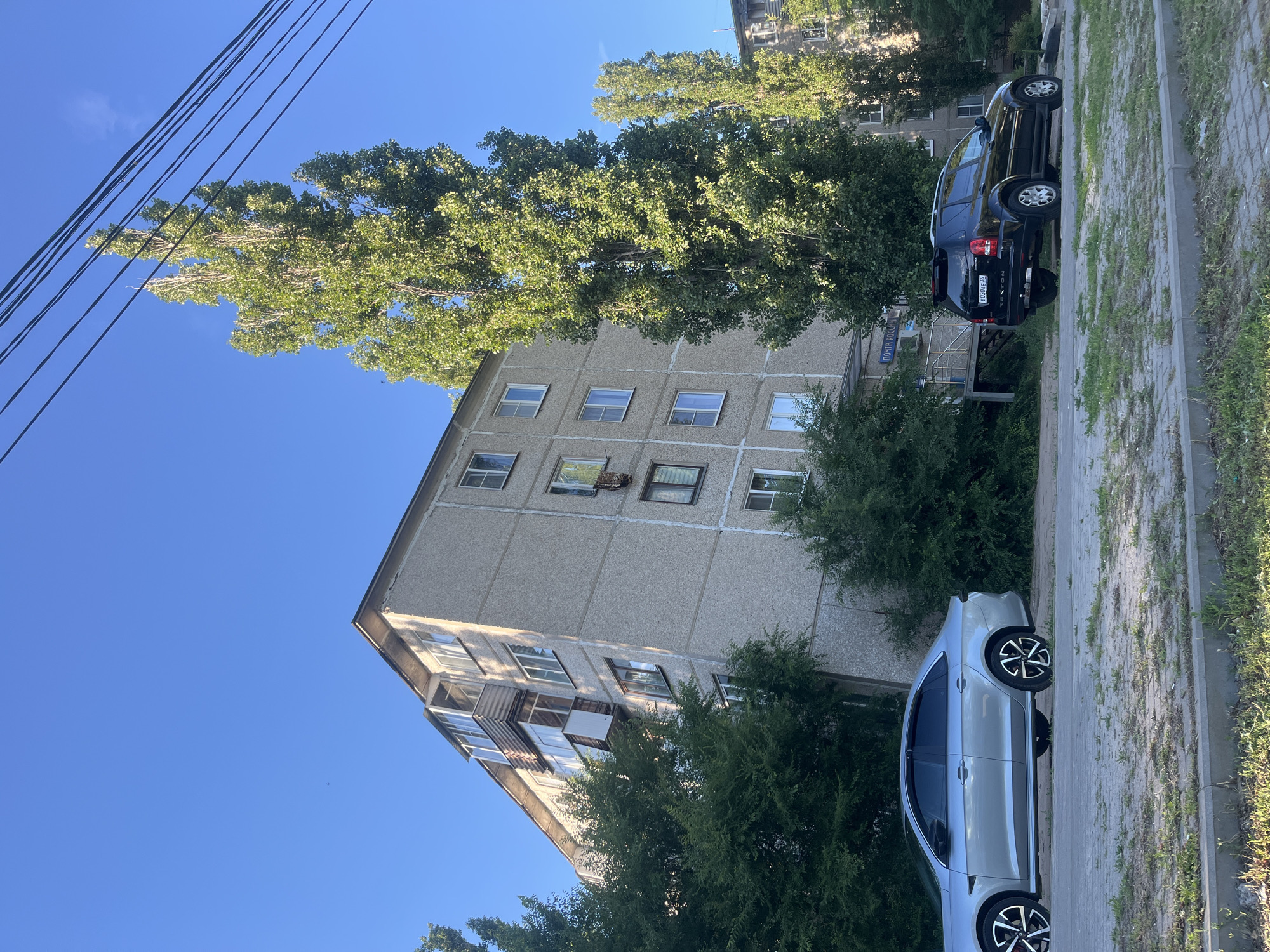 A silver sedan and a black SUV are parked on a paved street in front of a multistory apartment building. The building is gray with multiple windows and balconies. Tall, slender trees with green leaves are growing alongside the building. Overhead power lines are visible against a clear blue sky. The license plate on the black SUV reads "E 020 AB 36," suggesting it is registered in the Voronezh Oblast, Russia. A blue sign above the entrance to the building reads "ПОЧТА РОССИИ," indicating it is a Russian Post office. The surrounding environment appears to be an urban residential area.