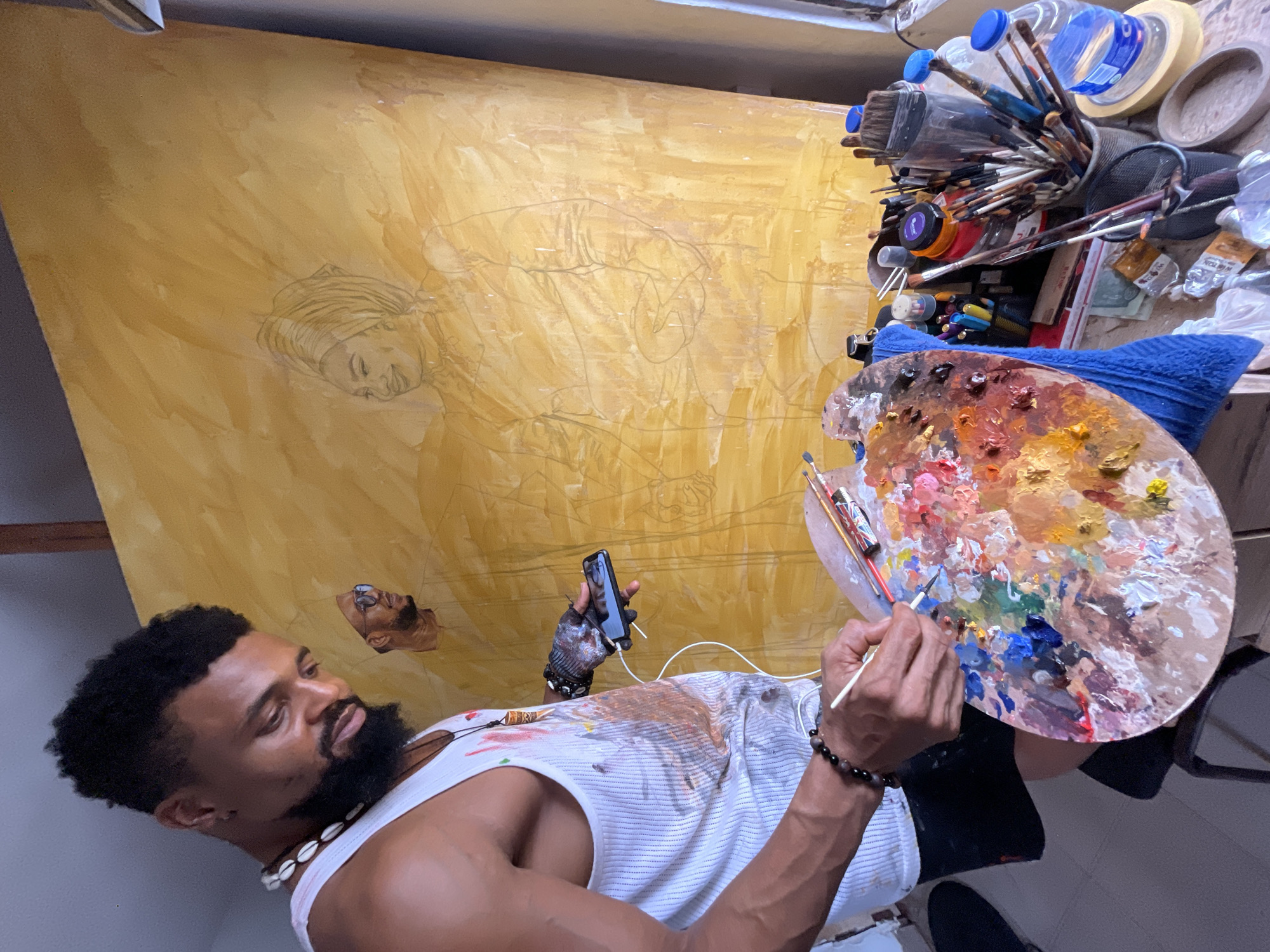 A medium shot shows a Black man in Ado, Nigeria, painting. He holds a brush in his right hand and a paint palette in his left. A large canvas behind him bears a sketch of a woman. A smartphone is held near his chest. He is wearing a white tank top with paint smudges and a shell necklace. Paint brushes and various art supplies are cluttered on a table to his right.