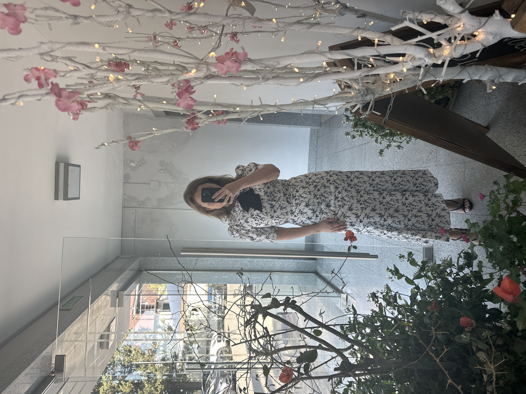 A woman is taking a selfie in a reflective glass-fronted entranceway, possibly a shop or hotel, with a narrow corridor visible behind her. She is wearing a white floral print dress with puffed sleeves and a black bow at the neck. She is holding a black smartphone in her right hand, obscuring her face. The environment features potted plants with red and pink flowers to the left, and a white decorative tree with pink blossoms and fairy lights to the right. A wooden A-frame sign is leaning against the decorative tree. In the background, through the glass, trees and buildings are visible, suggesting an urban setting. The floor is tiled. The lighting appears to be a mix of natural light from the entrance and artificial light from the ceiling and fairy lights. The location is identified as Sochi, Russia.