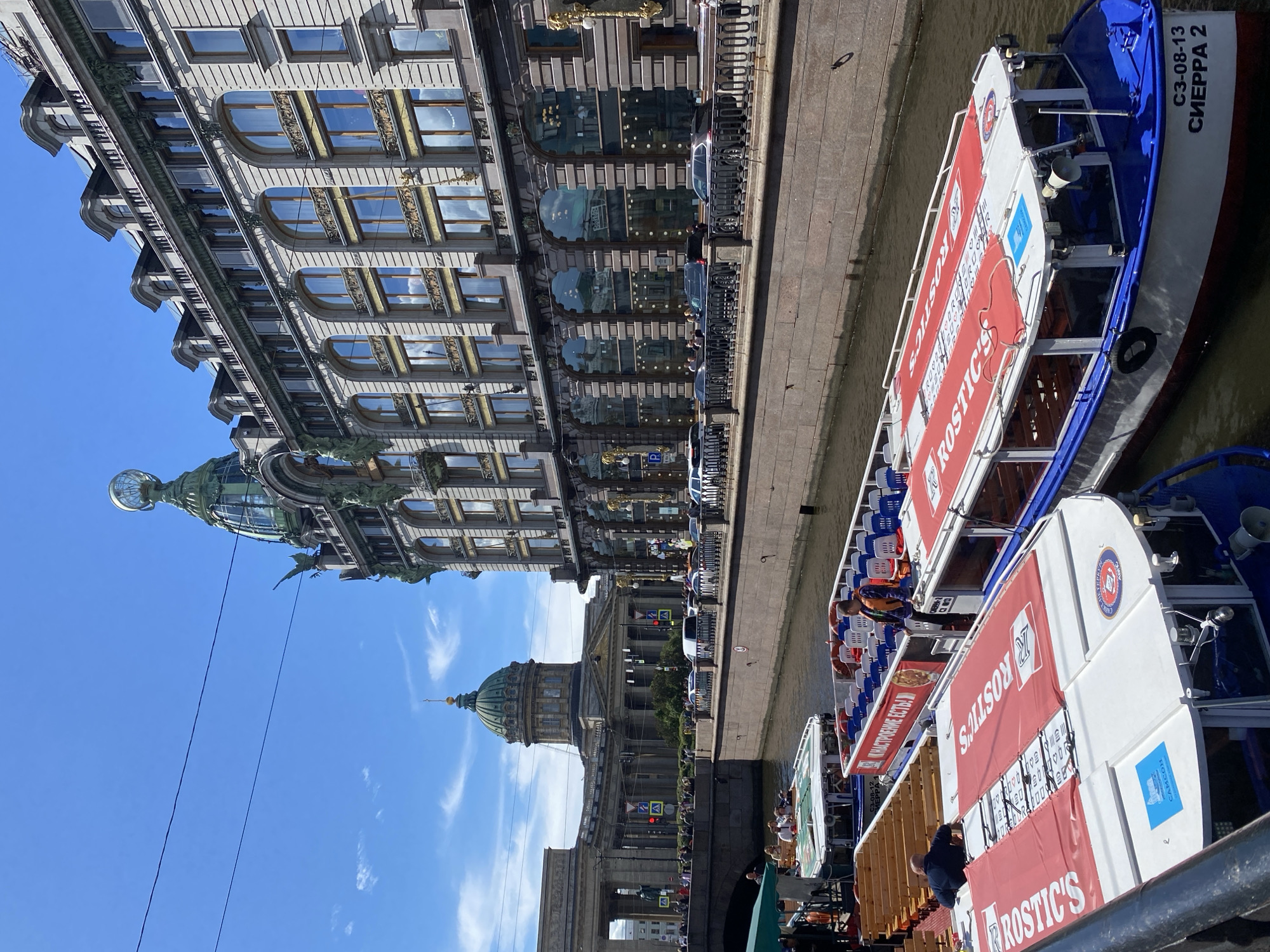 In Saint Petersburg, Russia, the Singer House, a landmark building with a distinctive green dome topped with a glass globe, stands prominently on Nevsky Prospekt. Its ornate facade, featuring numerous arched windows and gilded decorative elements, reflects the bright blue sky and scattered clouds. In front of the Singer House, a canal flows, with several tour boats docked along its edge. These boats, some with red awnings displaying the "ROSTIC'S" logo, are filled with passengers preparing for or disembarking from tours. The Kazan Cathedral, with its impressive colonnade and green dome, is visible in the background, adding to the grand architectural landscape of the city. The scene depicts a typical summer day in Saint Petersburg, with tourists enjoying the city's rich history and scenic waterways.