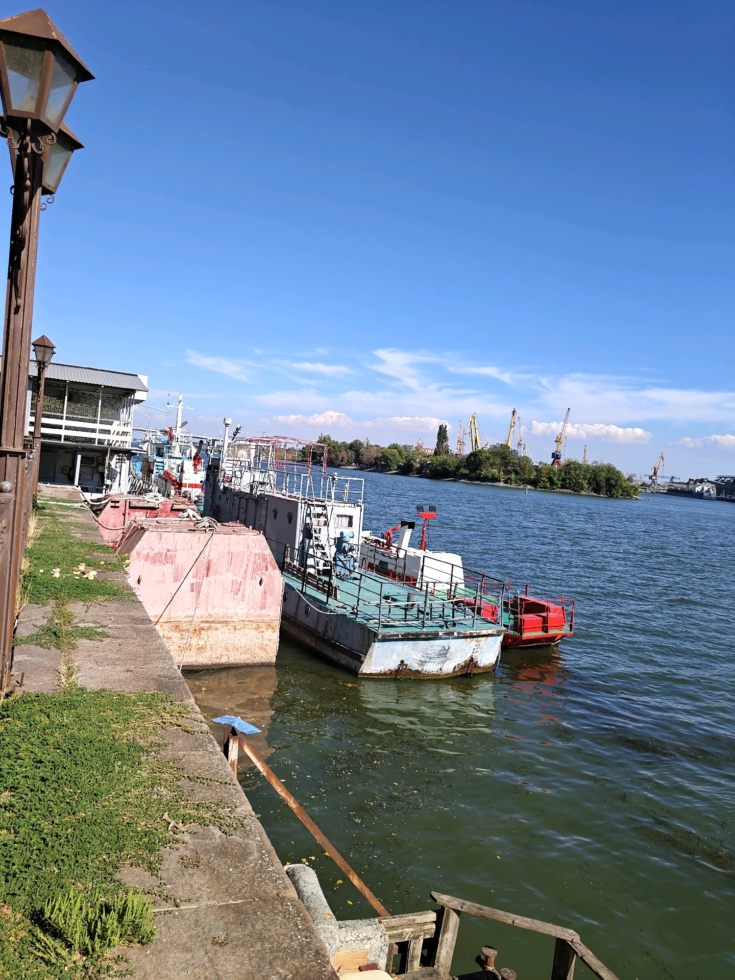 The image depicts a waterfront scene in the city of Rostov-on-Don, Russia, likely at a port or marina. In the foreground, a concrete embankment is visible, covered in patches of green grass and vegetation. A large, old-fashioned street lamp with two lights stands on the left side of the embankment.
Moored along the embankment are several boats of varying sizes and designs. One prominent vessel in the middle ground is a light blue and white boat with a red cabin and railing. To its left, a larger, rust-colored barge is docked. Further down the line, other boats with white superstructures and railings are visible.
The water in the foreground is a murky green, with visible debris floating on the surface. In the background, a wide body of water stretches out, with a distant shoreline dotted with trees and industrial cranes. The sky above is a clear, bright blue with scattered white clouds. The overall impression is one of a working port on a sunny day.