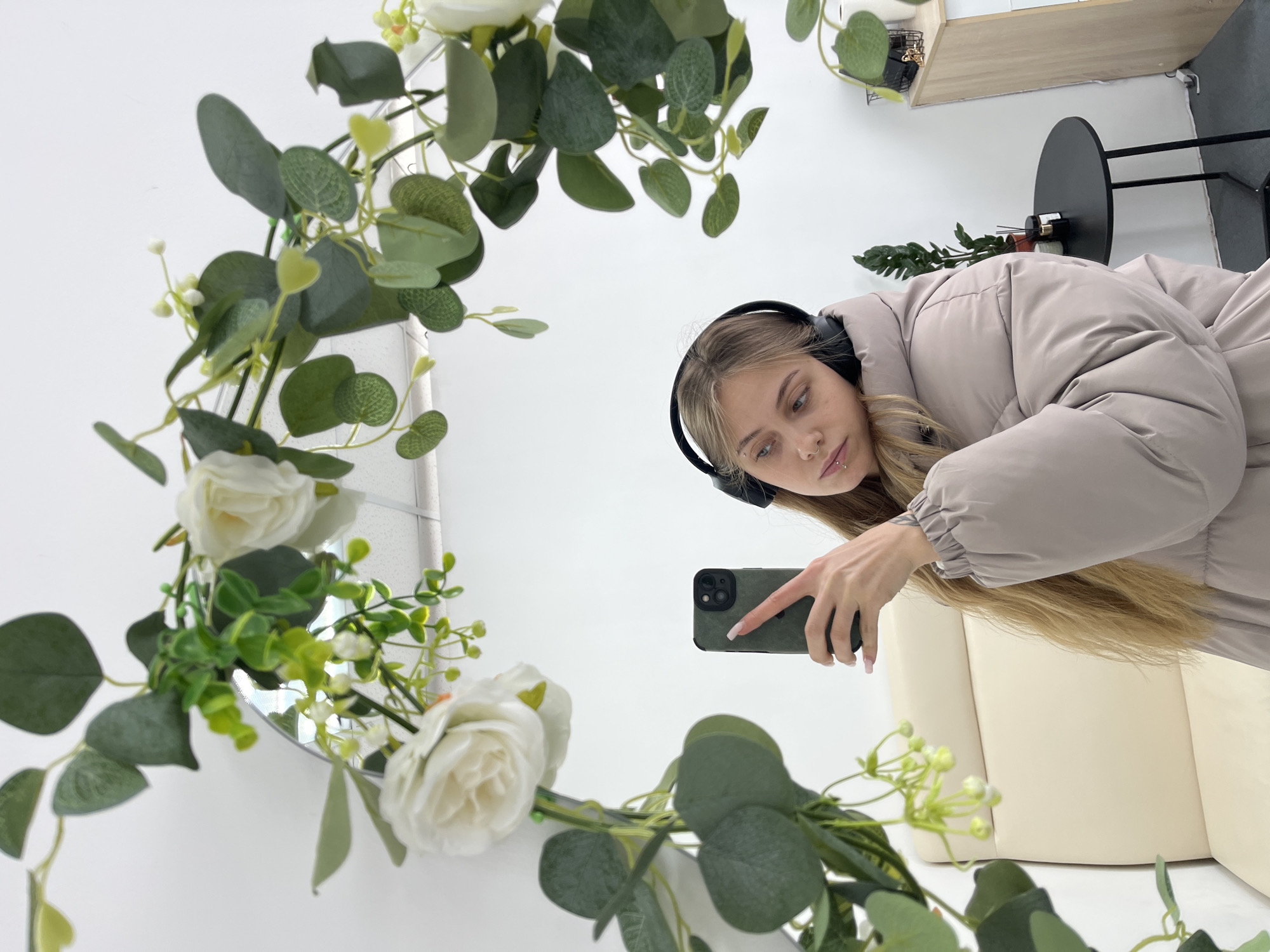 The image captures a mirror selfie taken by a young woman in an indoor setting. The central element is a large, possibly round or oval mirror, lavishly decorated with an artificial garland of green foliage and white flowers.
The woman, who appears to be in her late teens or early twenties, is positioned in the mirror's reflection. She has long, blonde hair that cascades over her right shoulder. She is wearing a light beige or tan puffer jacket with a slightly oversized fit and elasticized cuffs. Black over-ear headphones are worn on her head. Her face is directed towards the smartphone screen she is holding, with a focused and somewhat neutral expression. A small silver labret piercing is visible on her lower lip. Her right hand, with light-colored, manicured fingernails, holds a dark green smartphone, which is encased in a textured protective cover. The phone's camera module, featuring multiple lenses, is clearly visible on the back.
The artificial garland framing the mirror is dense with various types of green leaves, including larger, rounded ones resembling eucalyptus, and smaller, lighter green sprigs. Several white artificial roses, some fully bloomed and others in bud form, are interspersed throughout the foliage, along with clusters of tiny white buds and light green berries. This floral arrangement predominantly covers the upper and left portions of the mirror's frame.
The background reflected in the mirror reveals a clean, modern room. Behind the woman, a light cream or off-white upholstered sofa or armchair is visible. To the upper right of the reflected image (which would be to the left of the woman in reality), a small, round black side table with slender legs stands against a white wall. On the table, there's a dark object, possibly a diffuser or a decorative item, next to a small potted plant with dark green leaves. Above the table, a light-colored wooden shelf is mounted on the white wall, holding a few small, indistinct items. The floor visible in the reflection is light-colored, possibly white or light grey.
The lighting in the room is bright and even, suggesting either ample natural light or well-distributed artificial light, creating a clean and well-illuminated scene. The overall impression is that of a casual, self-portrait moment, highlighting both the individual and the decorated mirror.