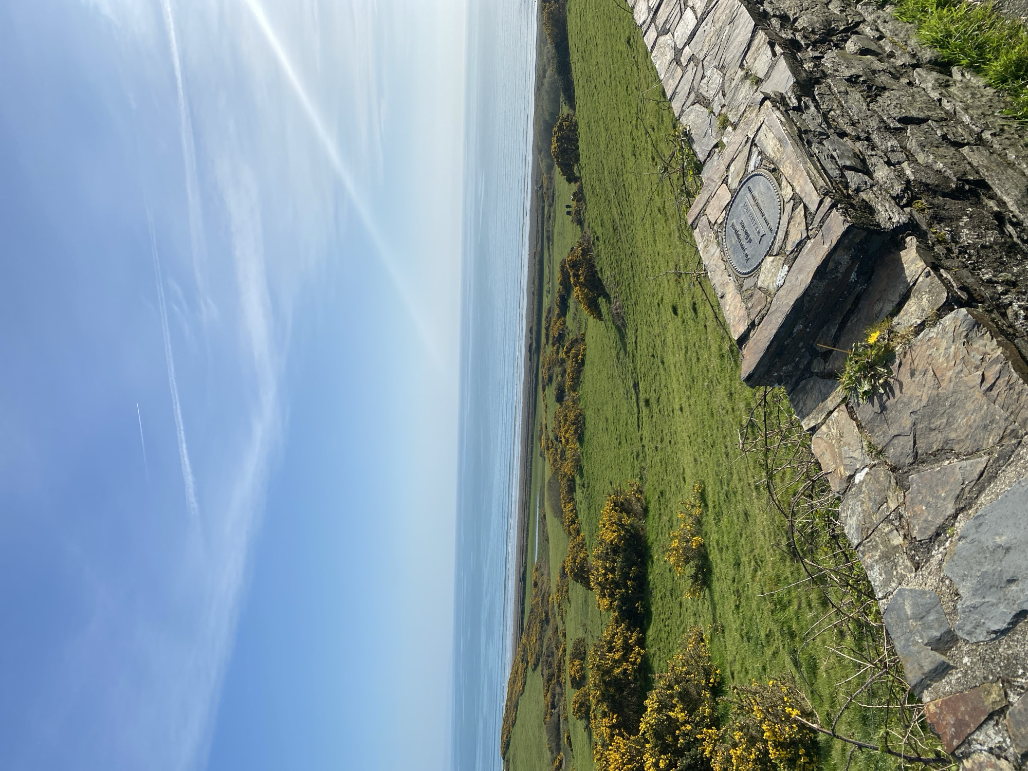 The image depicts a coastal scene in Criccieth, United Kingdom. The foreground shows a stone wall made of rough-hewn gray rocks. On top of one of the wall segments is a circular plaque. Beyond the wall, there is a green grassy field dotted with yellow flowering shrubs. The field extends to the edge of the sea, which is visible as a blue expanse. Above the sea, the sky is blue with streaks of white clouds and contrails from an aircraft. The image is taken from a high vantage point.