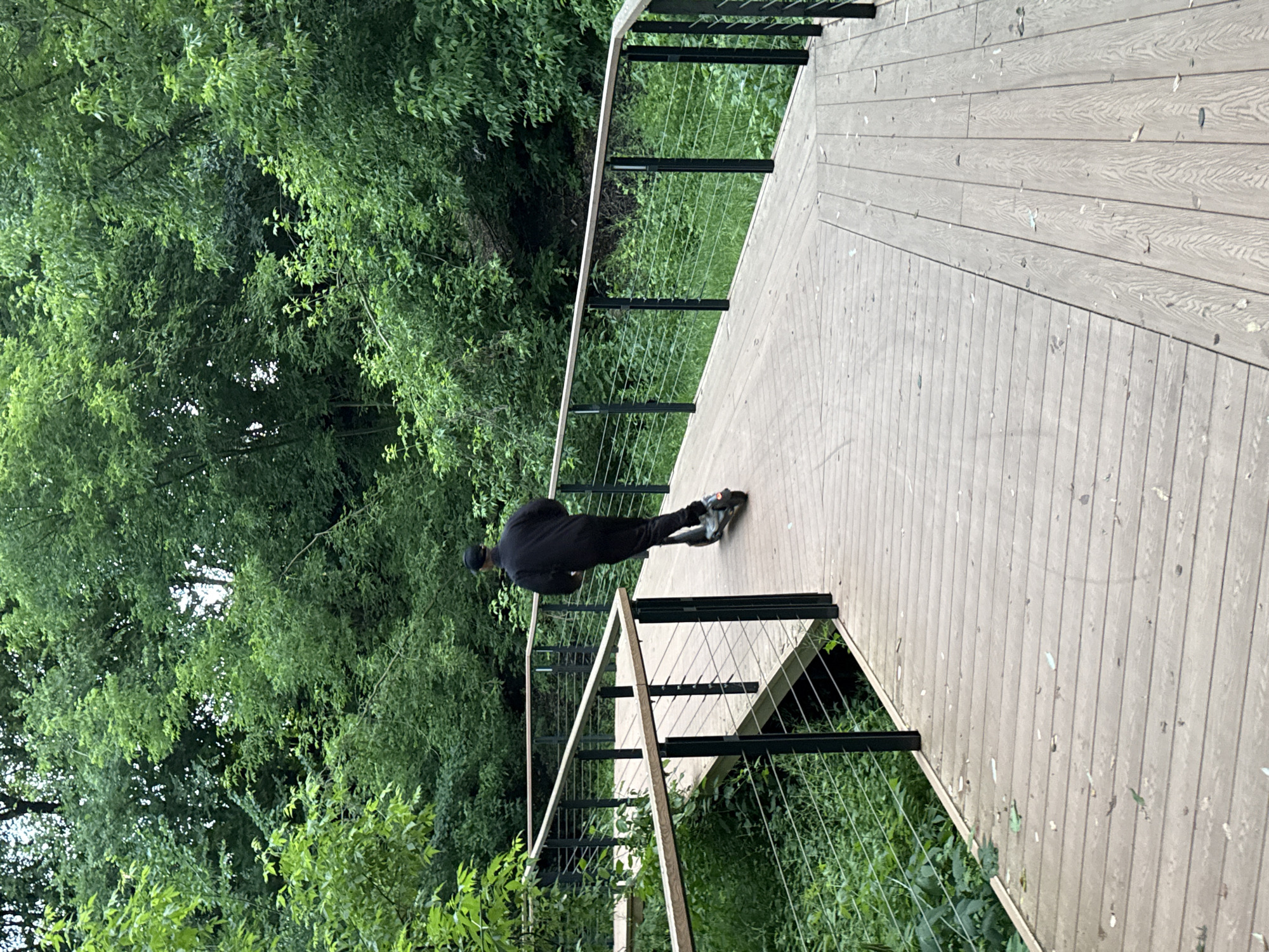 A person wearing a black tracksuit and a black baseball cap rides an electric scooter on a winding wooden boardwalk through a lush green forest. The boardwalk has dark metal railings with thin metal cables. The forest is dense with trees and undergrowth, suggesting a natural park or trail setting. The weather appears overcast. This scene is likely within the territory of Moscow, Russia, possibly in one of its large natural reserves or parks like Losiny Ostrov National Park or Bitsevsky Park, which are known for their extensive forest areas and well-maintained walking paths.