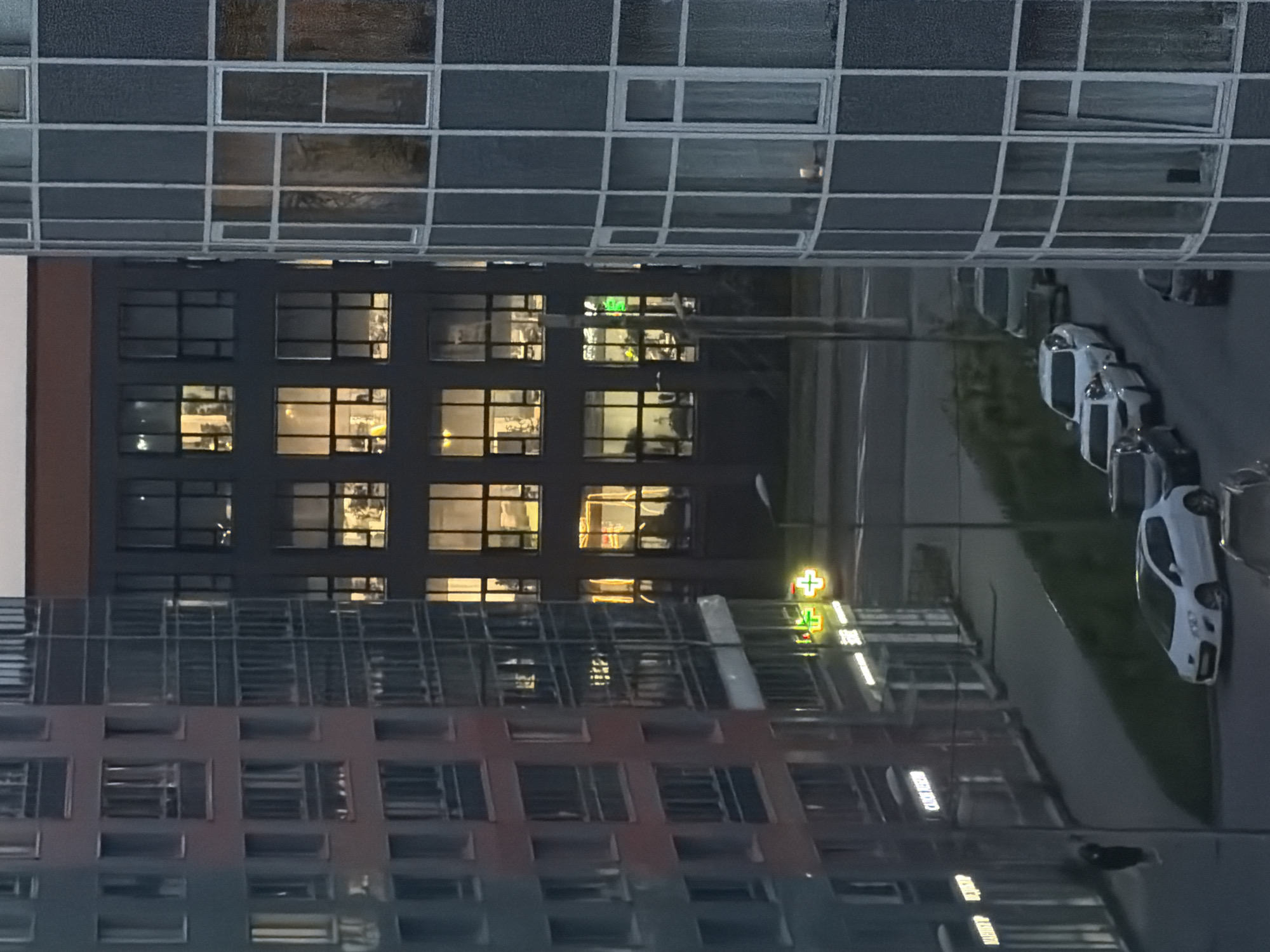This image, captured at dusk or nighttime from an elevated perspective, depicts a modern urban scene in Saint Petersburg, Russia. Three multi-story buildings define the view. On the left, a grey and reddish-hued residential or mixed-use building displays numerous windows and glass-fronted balconies. Its ground floor features commercial units with illuminated Cyrillic and Latin script signs; notably, a prominent green cross and a smaller white cross, typically signifying pharmacies, are visible. Other signs include "MARKET" and "CATCH ME IF YOU CAN." A single individual is discernible on the sidewalk adjacent to this building. To the right, a distinctively curved, multi-story building with a facade of glass panels and light grey frames stands prominently. In the background, a dark, multi-story structure, likely an office or commercial building, is brightly illuminated by numerous rectangular windows, revealing interior lighting. A paved street runs through the midground, occupied by several parked vehicles, including a white hatchback and multiple darker sedans. The scene is predominantly lit by artificial light sources from the buildings and street-level signage, consistent with contemporary urban development in Saint Petersburg.
