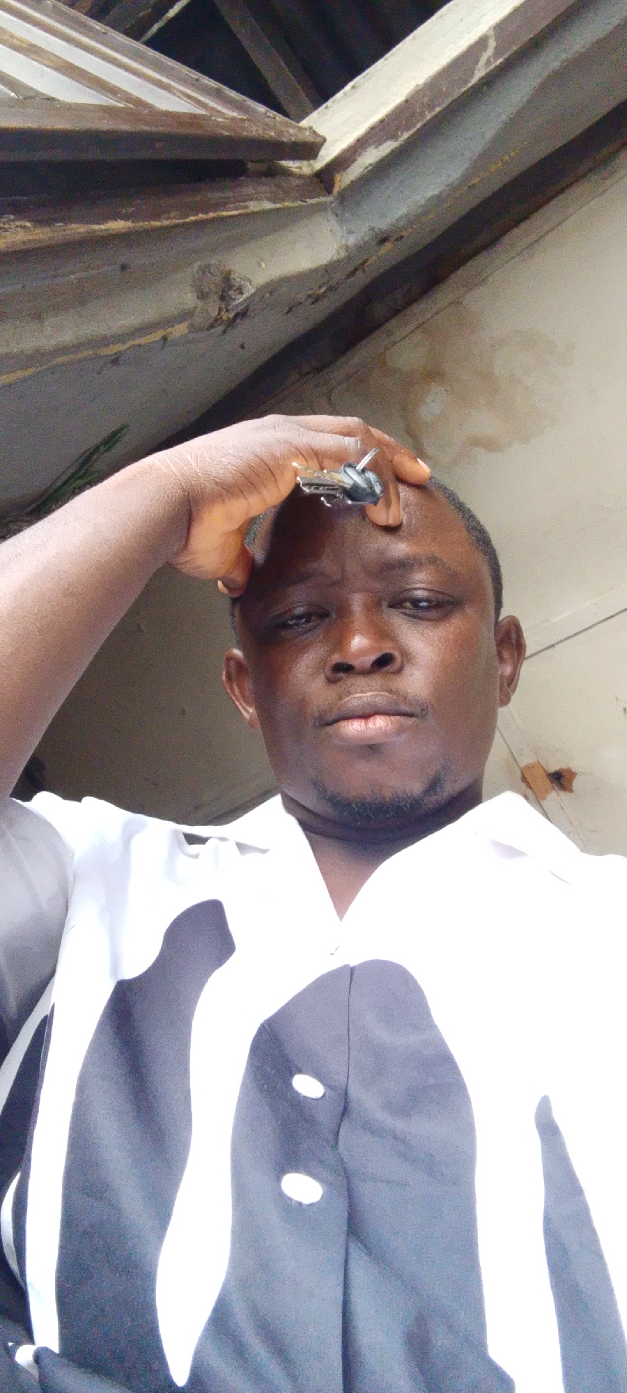 A man is holding keys to his forehead inside a structure with exposed wooden beams. The man is dark-skinned, wearing a white and black shirt, and looking directly at the camera. The location is Lokoja, Nigeria.