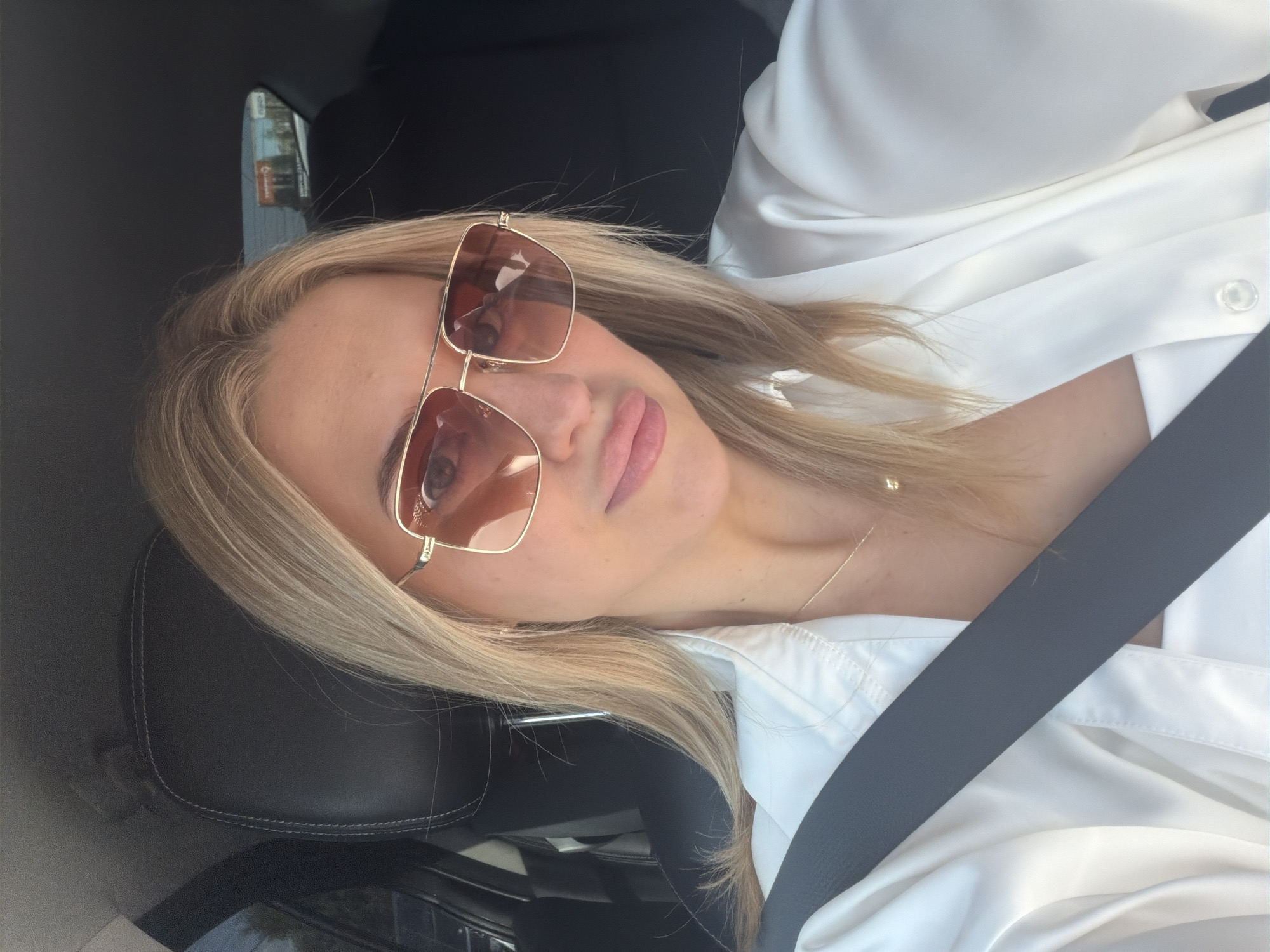 A woman with blonde hair, wearing sunglasses and a white shirt, is inside a car. She is sitting in the driver's seat, with a seatbelt across her body. The car interior is visible, including a black leather headrest and part of the dashboard. The woman is looking at the camera and has her lips slightly parted. The scene is captured during the daytime, with natural light illuminating the woman's face and clothing. There are no overt indicators of the specific location within Kyiv, Ukraine, beyond it being an urban environment as suggested by the background glimpse of a billboard.