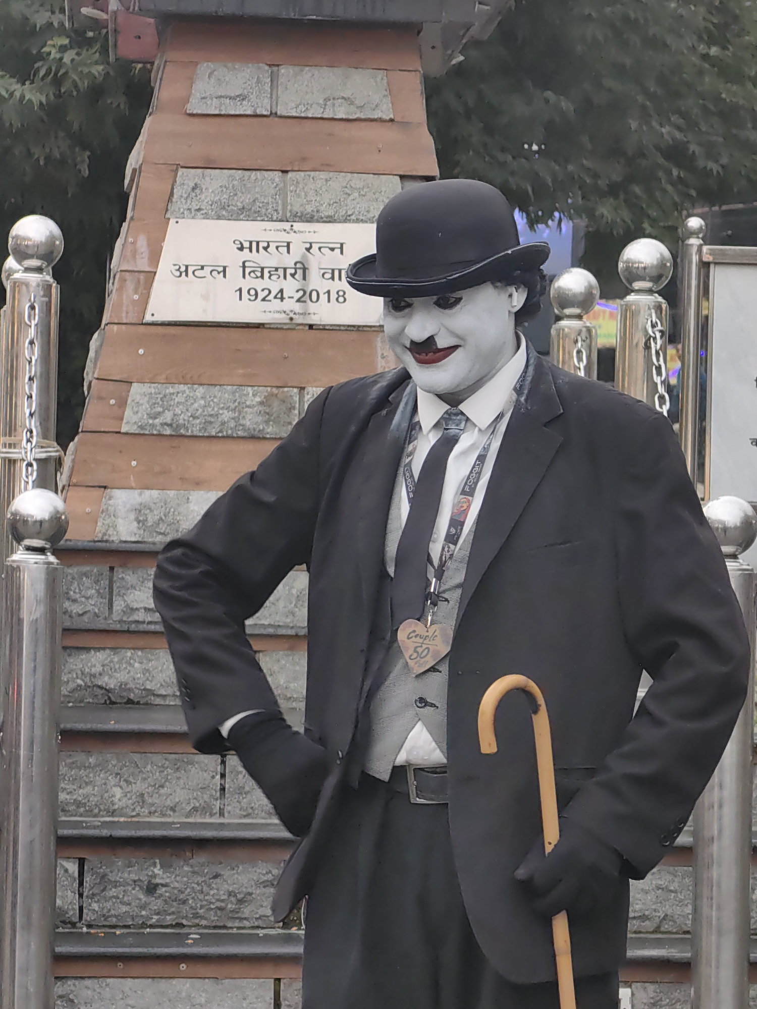 A person dressed as Charlie Chaplin, with white face paint, black lipstick, and a black bowler hat, stands on stone steps in front of a wooden structure with a plaque. The plaque, in Hindi, reads "Bharat Ratna Atal Bihari Vajpayee 1924-2018". The person is wearing a black suit, a grey vest, a white shirt, a black tie, black gloves, and holds a light brown cane. A heart-shaped tag reading "Couple 50" is attached to a lanyard around their neck. The background shows trees and what appears to be a chain fence. This scene is located at the Atal Bihari Vajpayee memorial in Manali, India.
