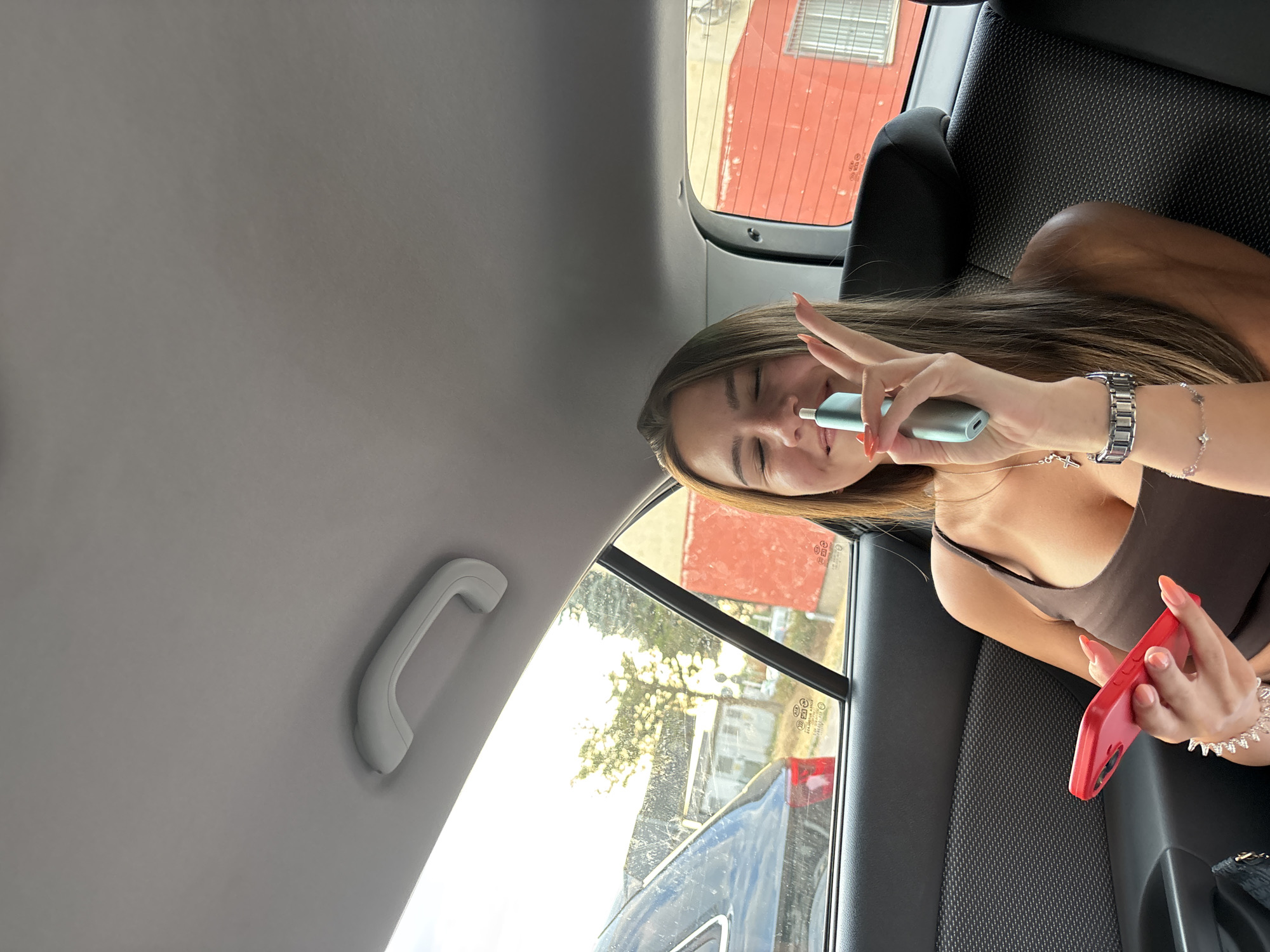 The image shows a young woman sitting in the back seat of a car. She is holding a red smartphone in her left hand and a light blue electronic cigarette in her right hand, which she is bringing to her face. She is wearing a brown tank top and a silver necklace with a cross pendant. She has long, brown hair and painted orange fingernails. A silver bracelet adorns her left wrist, and a watch is on her right wrist. Through the car window, a blurry background of buildings and trees is visible, with the sun setting in the distance. The car appears to be in motion, indicated by the blur in the background. The setting is likely a street or road within the city of Vratsa, Bulgaria, given the presence of buildings and a typical urban environment, possibly in the late afternoon or early evening due to the lighting conditions.