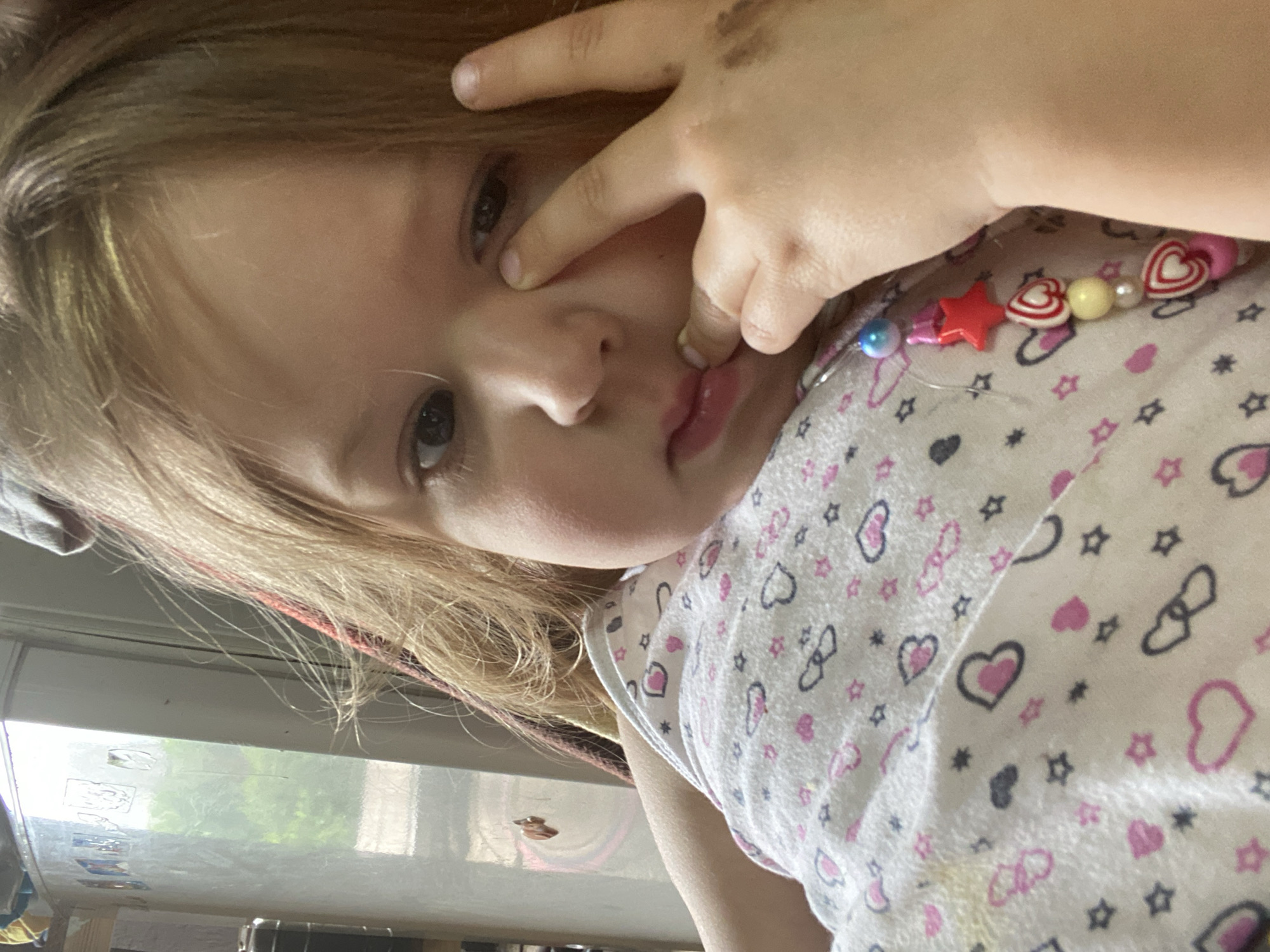 The image shows a close-up of a young child's face and upper body. The child has fair hair and appears to be looking down, with a finger near their eye. They are wearing a white top with a pattern of pink and dark blue hearts and stars. A colorful beaded necklace with red stars and heart-shaped beads is visible on the child's chest. The background is out of focus but suggests an indoor setting with a window and what appears to be a refrigerator to the left. The image does not provide any specific information about the city of Chaikovsky, Russia.