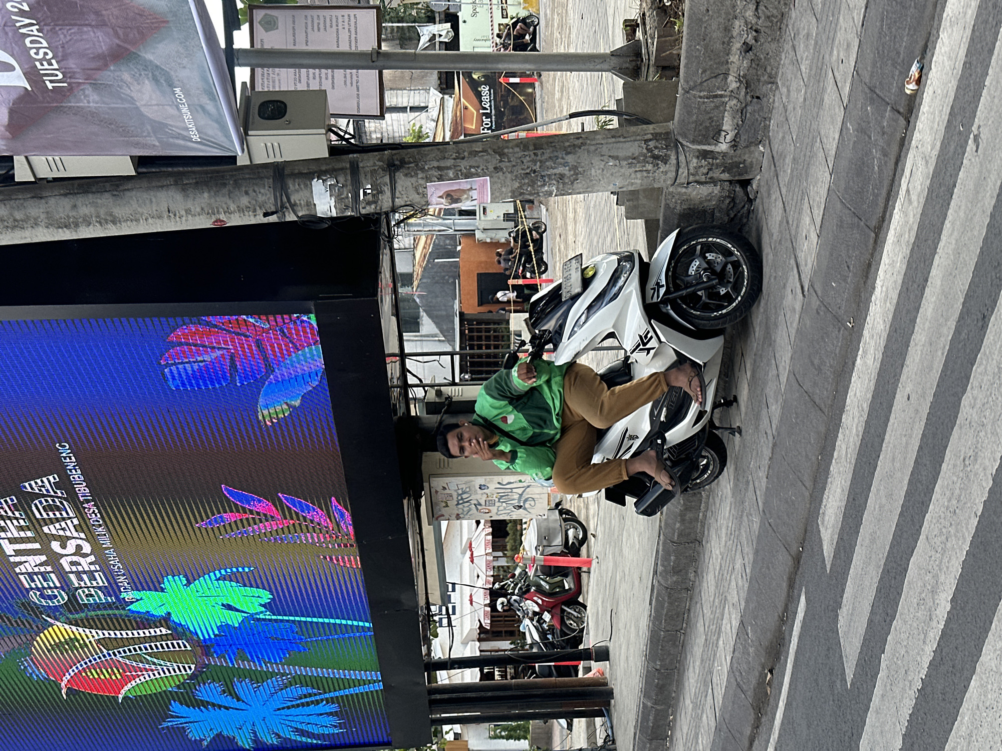 A man sits on a white scooter on a sidewalk in front of a large digital billboard in North Kuta, Indonesia. He is wearing a green jacket and brown pants, with earbuds in his ears. He is smoking a cigarette and looking to his left. The billboard displays colorful graphics and text, including "GENTRA PERSADA" and "SELASA 25". Behind the man, several other scooters are parked, and buildings and shops are visible. The sidewalk is paved with concrete and has a curb separating it from the street. A pedestrian crossing is marked on the street in front of the sidewalk. The environment appears to be a busy urban area, with streetlights and electrical wires visible.