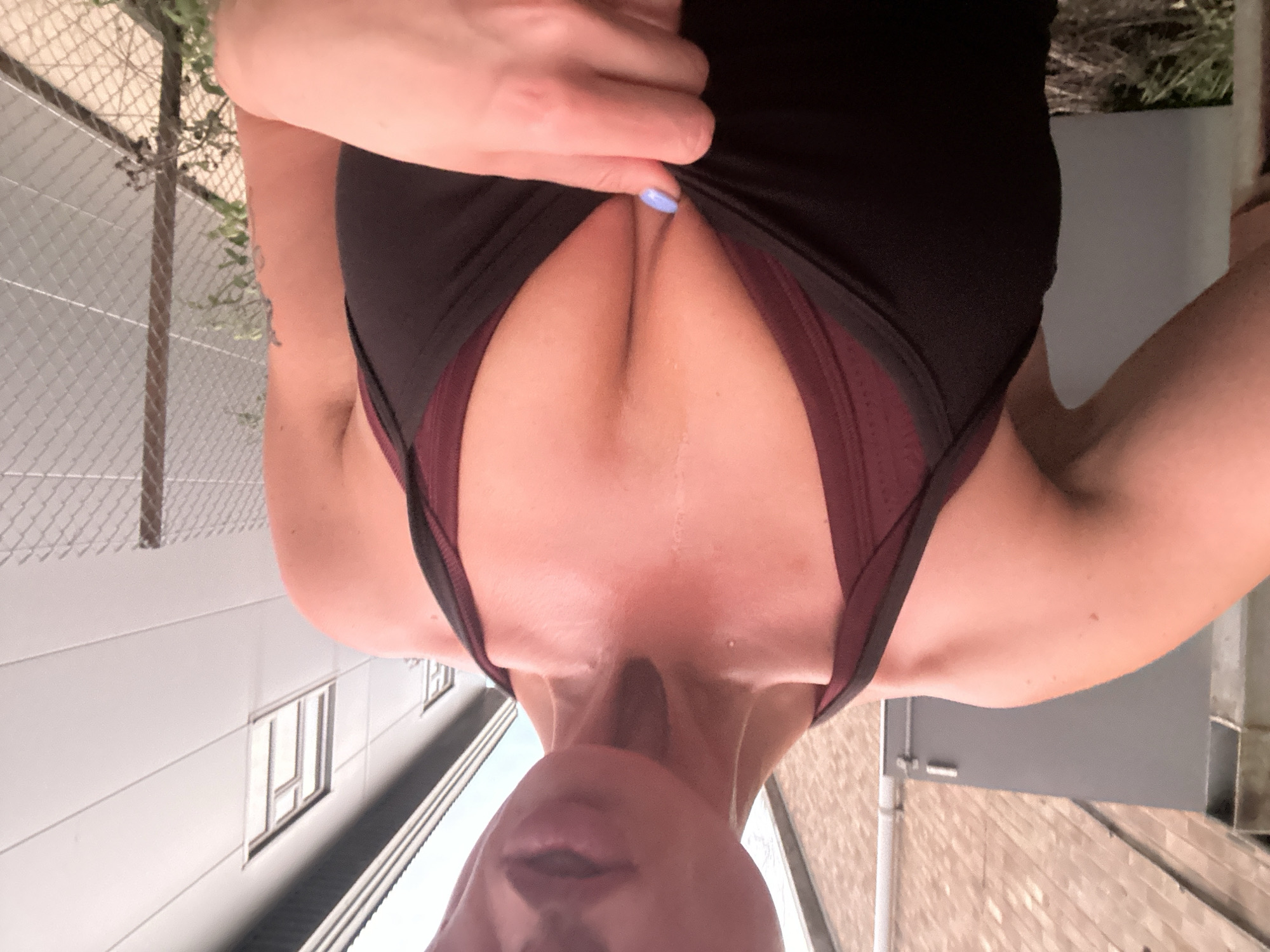 The image presents an inverted close-up view of a person from the chest upwards. The individual wears a black tank top over a dark red sports bra. A hand, adorned with light blue nail polish, pulls at the black top, emphasizing the cleavage. The subject's chin, mouth, and nose are visible at the bottom of the frame, suggesting a backward head tilt.
The outdoor background, when oriented correctly, shows a chain-link fence with some green foliage on the left. Behind the fence, a light-colored building with rectangular windows is visible. On the right, a gray building wall, possibly with a downspout, and light brickwork are present. The bright lighting indicates daytime. No text is visible. This scene is located in Tonbridge and Malling, United Kingdom.