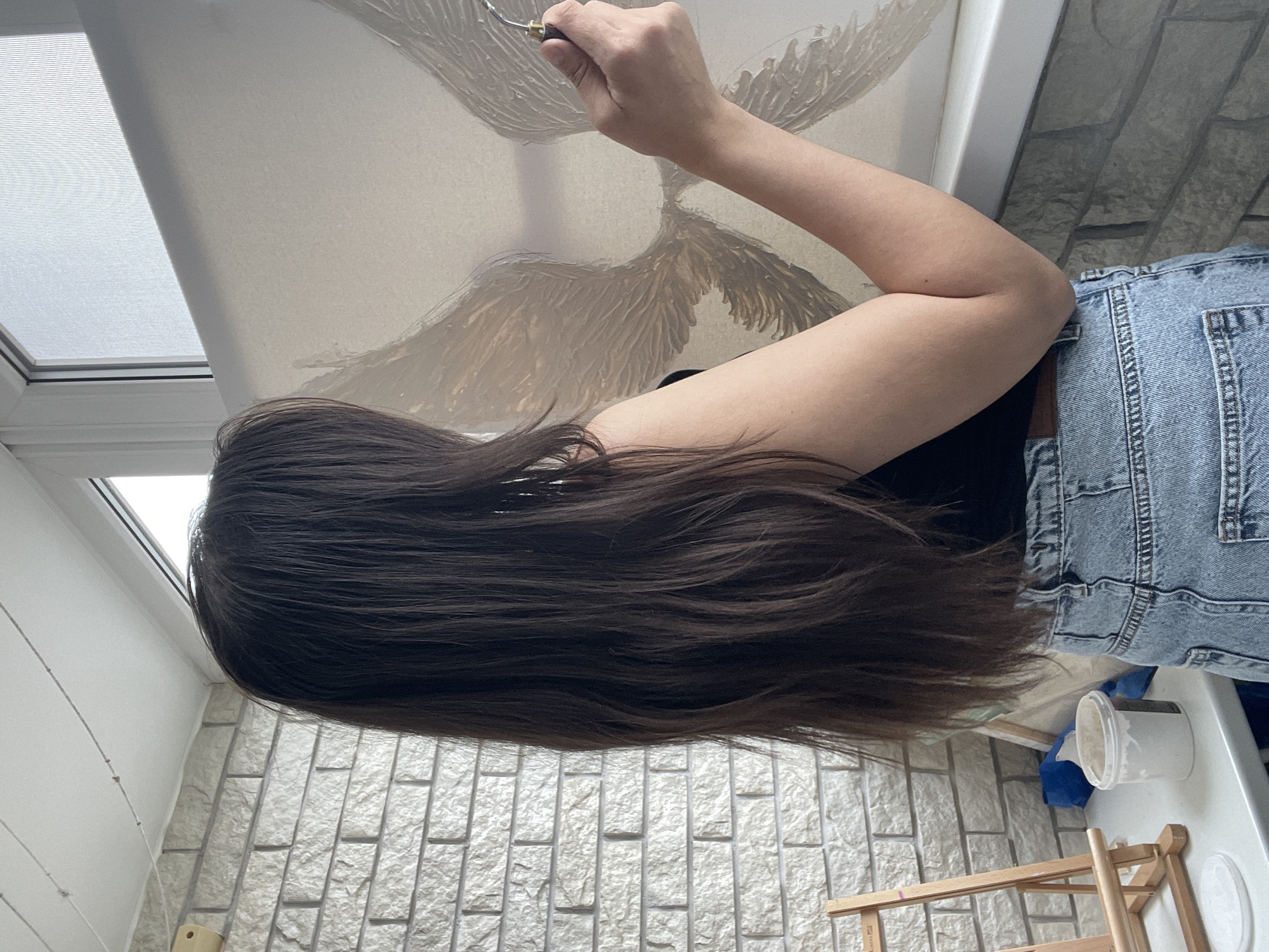 A person with long, dark hair is painting on a canvas attached to a frame. The person is wearing a black shirt and light-blue denim shorts. They are using a palette knife to apply paint in a wing-like pattern. The scene is indoors, with a window at the top left. The room has textured light-grey walls. A white table is visible at the bottom right, with a container and some wooden objects on it. The location appears to be a studio or art space in Unknown, Unknown.