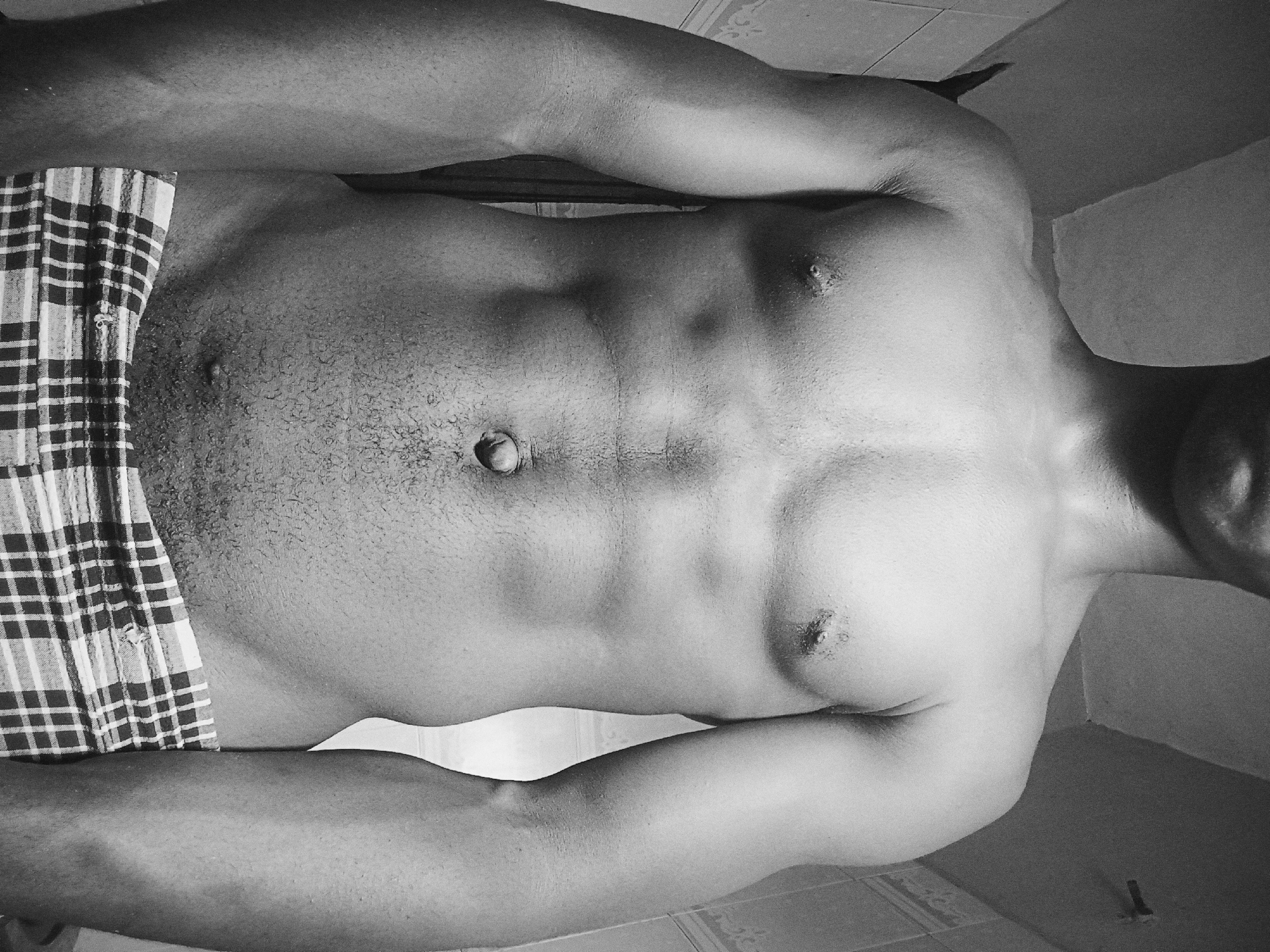 A black and white image depicts a shirtless man wearing plaid shorts, his torso and upper body prominently displayed. He is standing in a bathroom; tiles are visible on the floor and wall. The location is Nkpor, Nigeria.