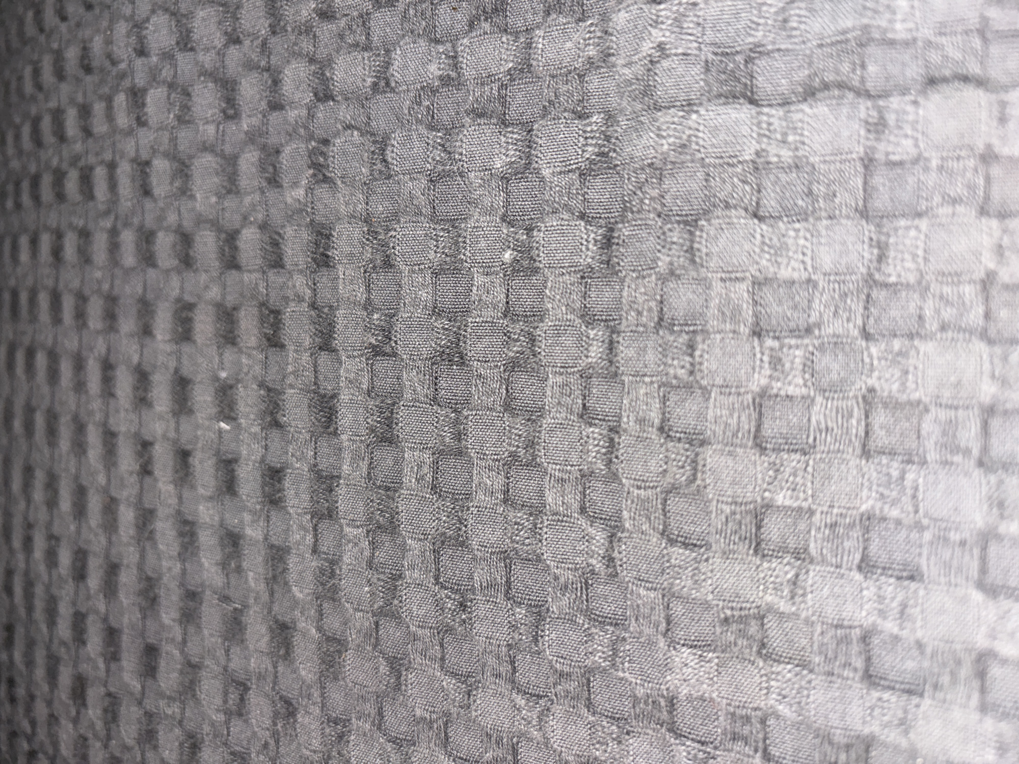 The image is a close-up, angled shot of a grey, woven fabric with a raised, checkered pattern. The texture of the fabric appears to be somewhat rough or nubby, with individual threads visible within the pattern. The lighting creates shadows within the crevices of the weave, emphasizing the three-dimensional quality of the pattern. The perspective suggests the fabric is laid out flat, possibly on a surface, and the focus shifts from sharp in the foreground to slightly blurred in the background, giving a sense of depth. There are no people, objects, or environmental features that would indicate a specific location in Cupra Marittima, Italy.