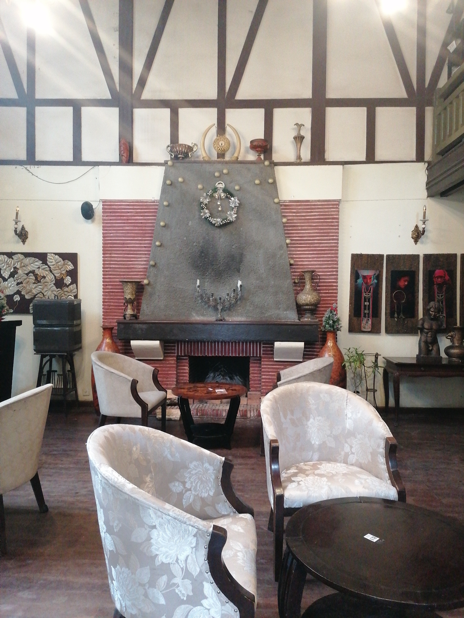 This media file captures the interior of a cozy, traditionally decorated lounge or common area, likely within a hotel or cafe in Da Lat, Vietnam. The setting is characterized by a prominent Tudor-style aesthetic, featuring dark timber beams and white plaster infill on the upper walls and ceiling.
The central focus is a large, dark grey concrete or stone fireplace, adorned with a festive silver and white wreath. The fireplace is flanked by walls of red brick. On its mantelpiece, various decorative items are displayed, including metallic urns, an ornate candelabra, and tusk-like sculptures.
The room is furnished with several light-colored armchairs, upholstered in a cream or beige fabric with a subtle floral pattern. These chairs have dark wooden frames and are arranged around small, dark, round wooden coffee tables.
Decorative elements are abundant throughout the space. On the left wall, a large artwork with raised botanical motifs in silver and grey is mounted, with what appears to be stacked audio equipment or boxes on a stand below it. A wall sconce with a candle provides ambient light. On the right wall, three vertically oriented artworks depict stylized portraits, possibly of local or indigenous figures. Below these, a classical male torso sculpture and several decorative vases sit on a dark wooden table. Another wall sconce is visible on this side.
No people are visible, suggesting a quiet atmosphere. The lighting is dim but with a bright glare in the upper left, possibly from a window or skylight, indicating it might be daytime. A small white sticker, possibly a price tag, displaying "89" is faintly visible on one of the tables in the foreground. The overall ambiance is one of comfortable elegance with a strong traditional and cultural flair.