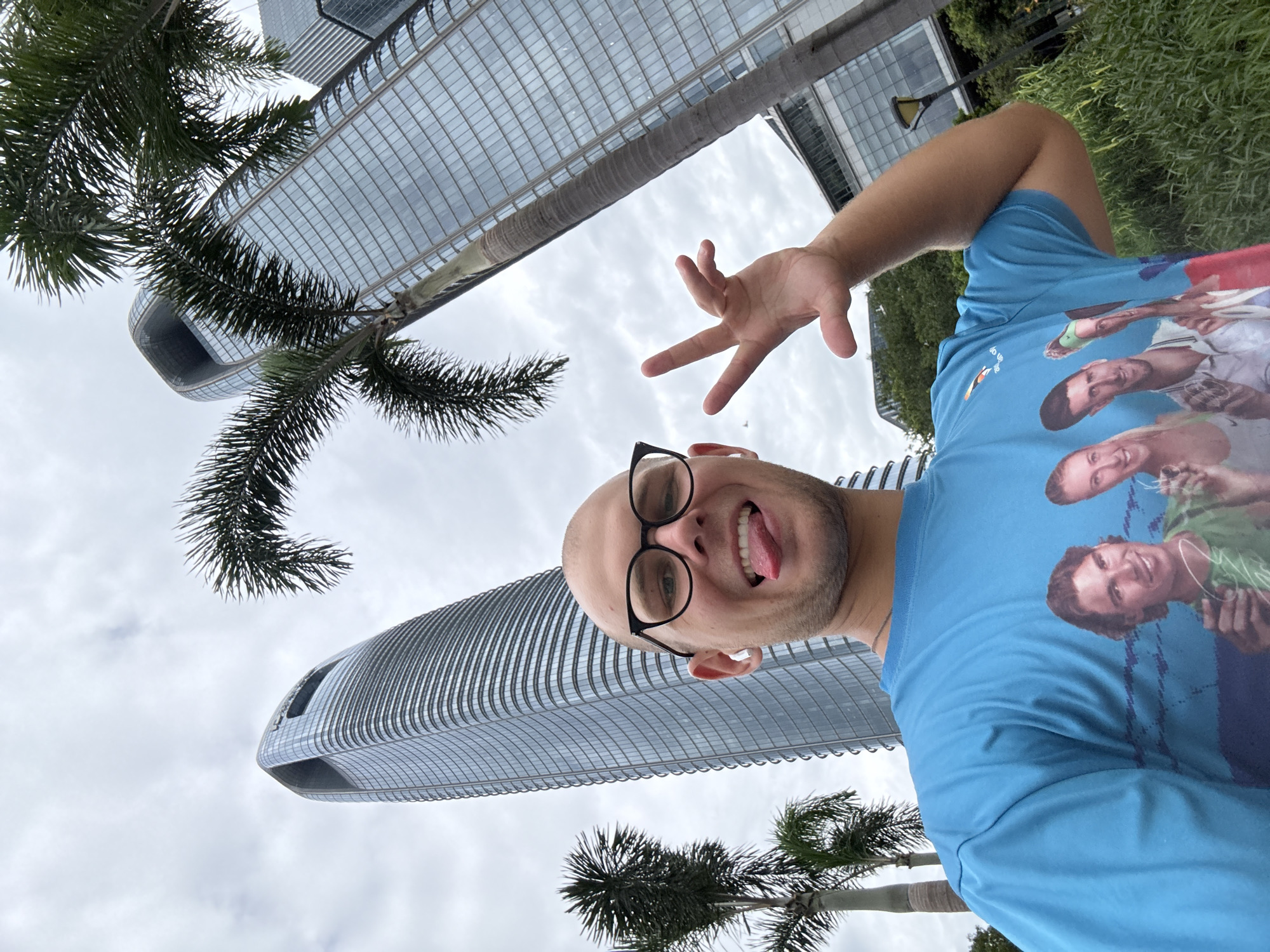 A man with a shaved head, wearing glasses and a blue t-shirt, sticks his tongue out at the camera while making a hand gesture with his right hand. His t-shirt features a graphic of four tennis players, including Rafael Nadal and Novak Djokovic. He is standing in front of two modern skyscrapers and palm trees. The scene appears to be in the Tianhe District of Guangzhou, China, as indicated by the architecture of the buildings and the presence of palm trees. The sky is overcast.