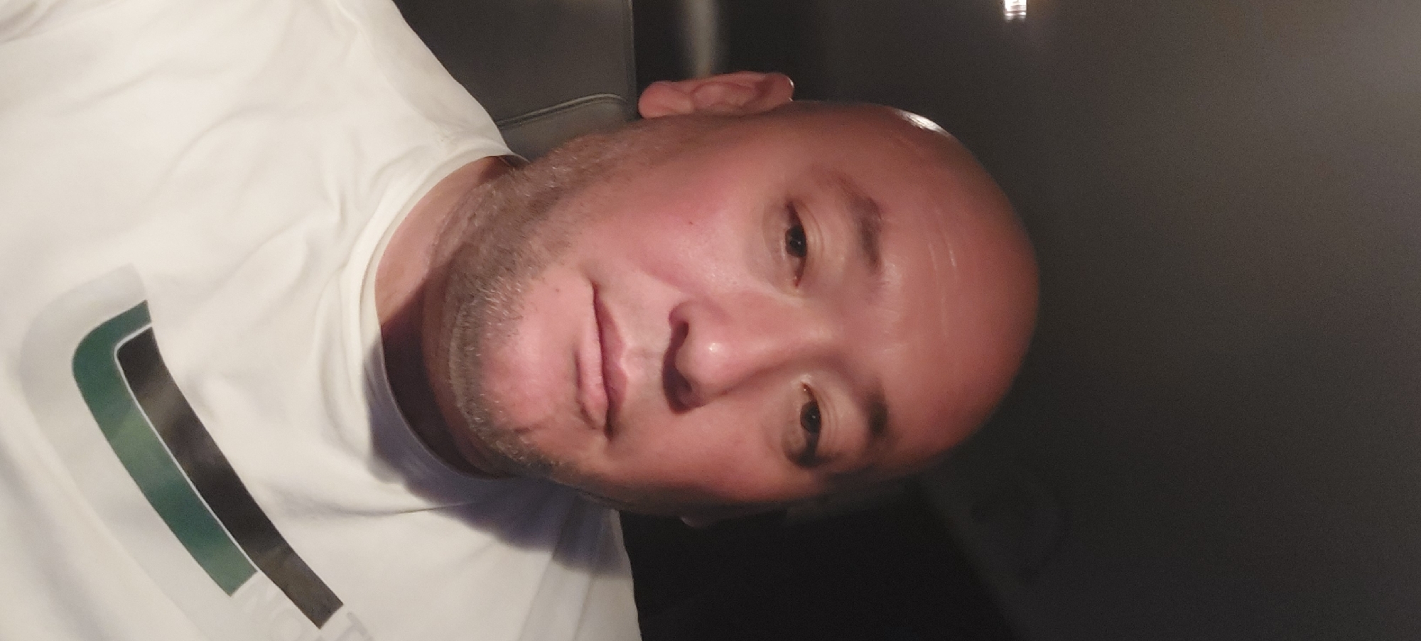 A close-up, head-and-shoulders selfie taken from a low angle shows a bald, middle-aged man with fair skin and light stubble on his face. He is wearing a white t-shirt with a graphic design on the chest. The background is dark and appears to be the interior of a vehicle, with a dark seat visible behind the man's left shoulder. The lighting suggests an indoor or nighttime setting. No specific location in Fergana City, Uzbekistan is discernible from the image.